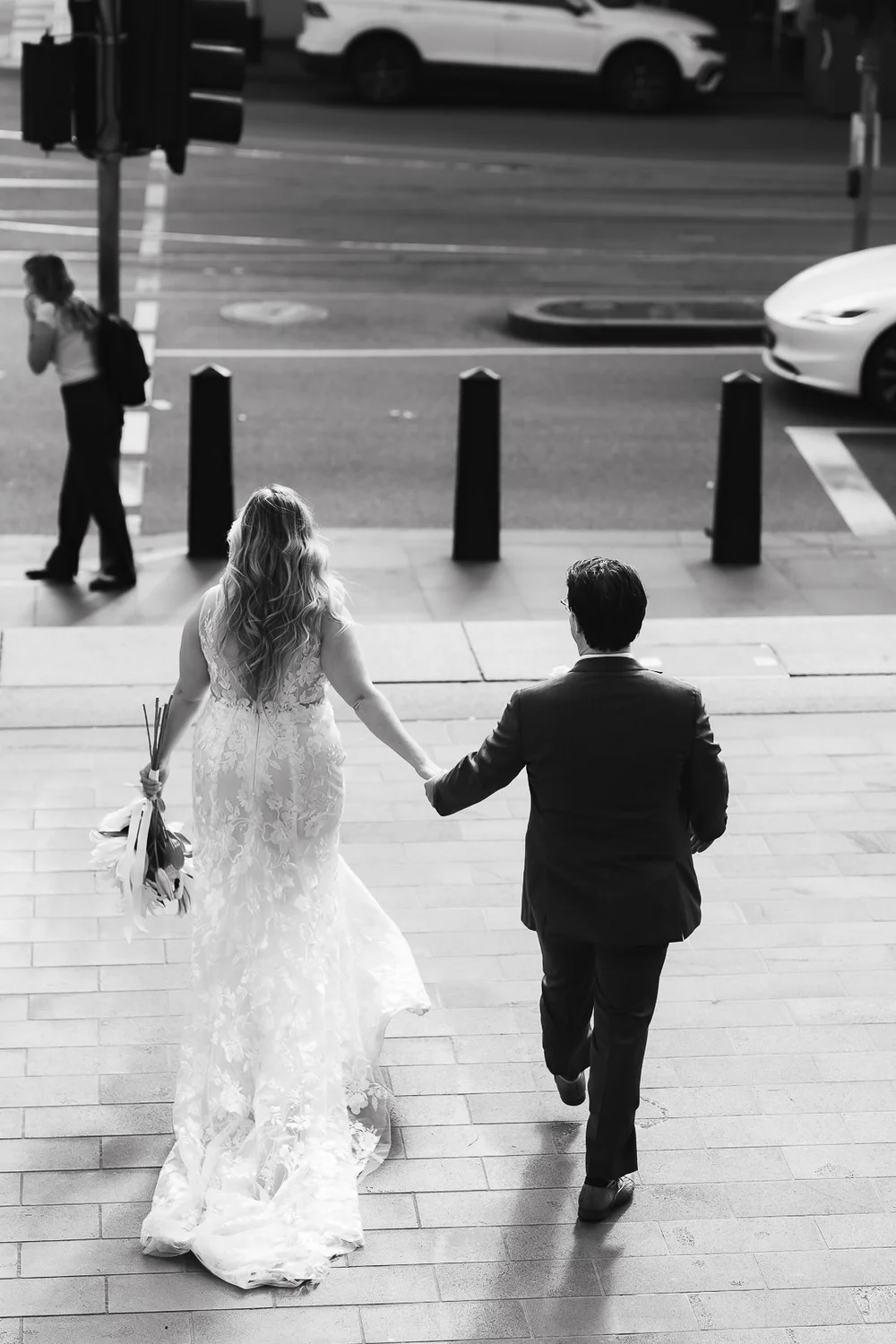 A bride and groom holding hands, walking up steps outside a building, with a city street in the background.