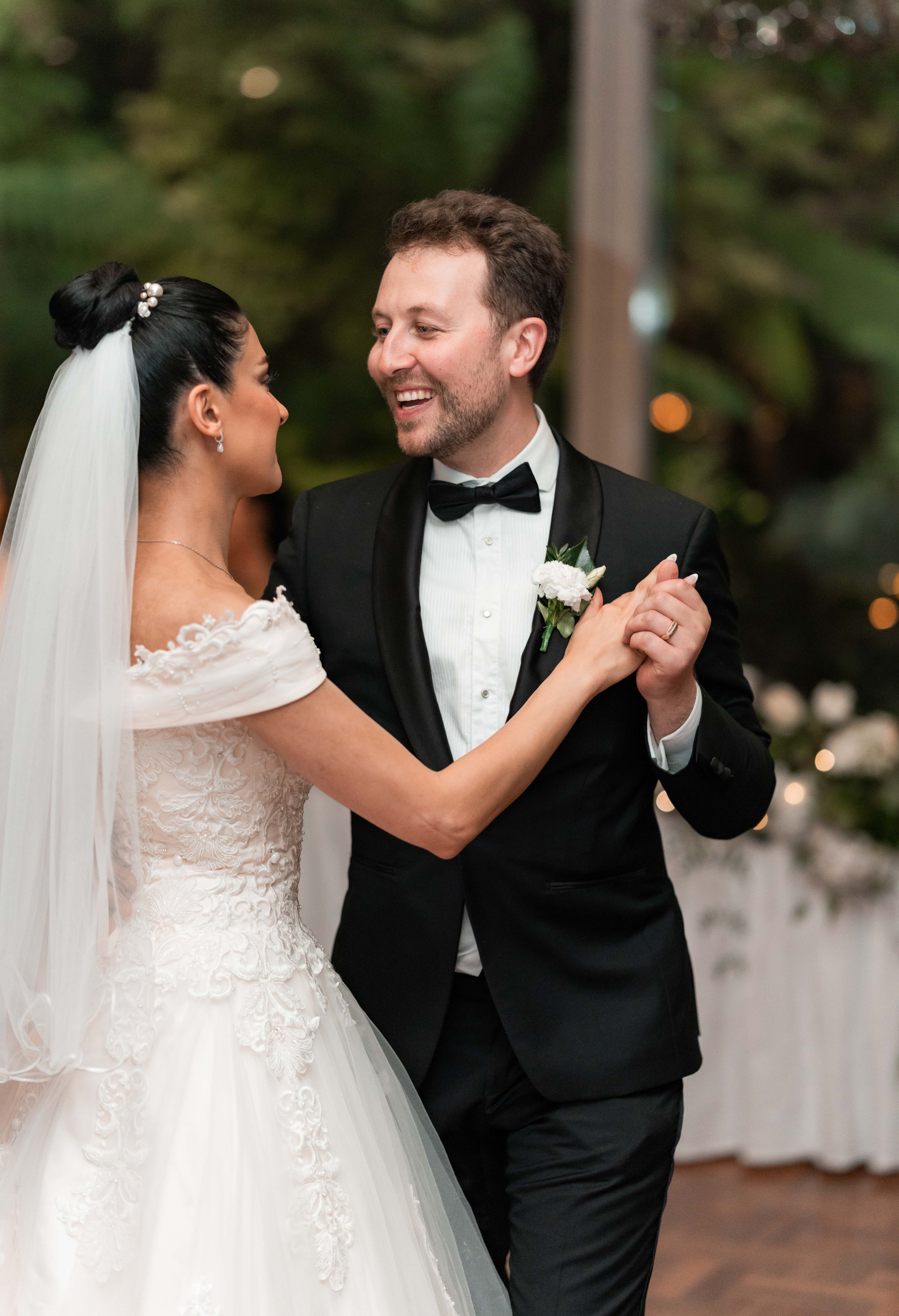 Bride and groom dancing at wedding reception, smiling at each other, with trees and decorations in the background.