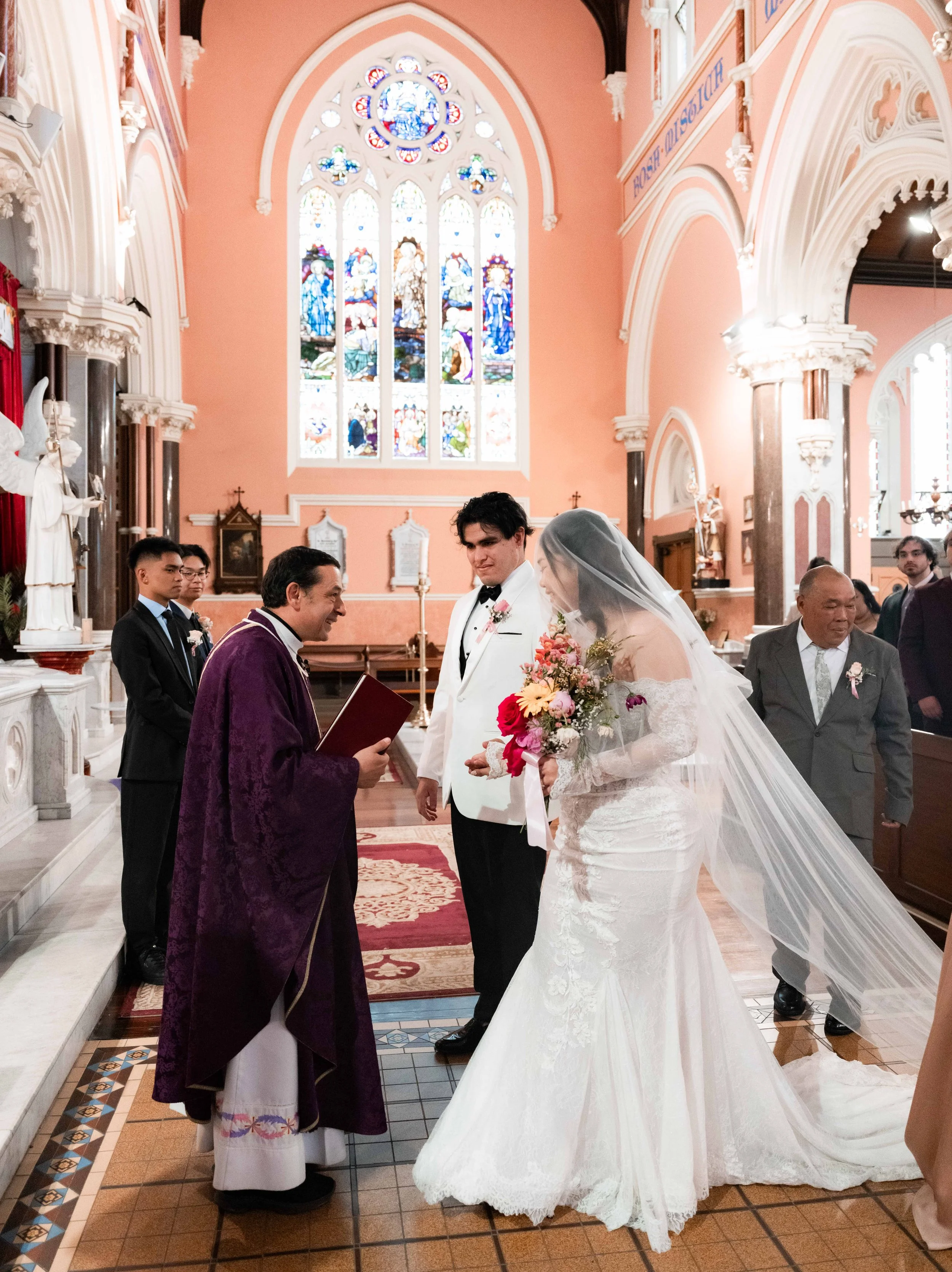 A wedding ceremony inside a church with a stained glass window in the background. The bride and groom are at the altar with a priest, and guests seated nearby.