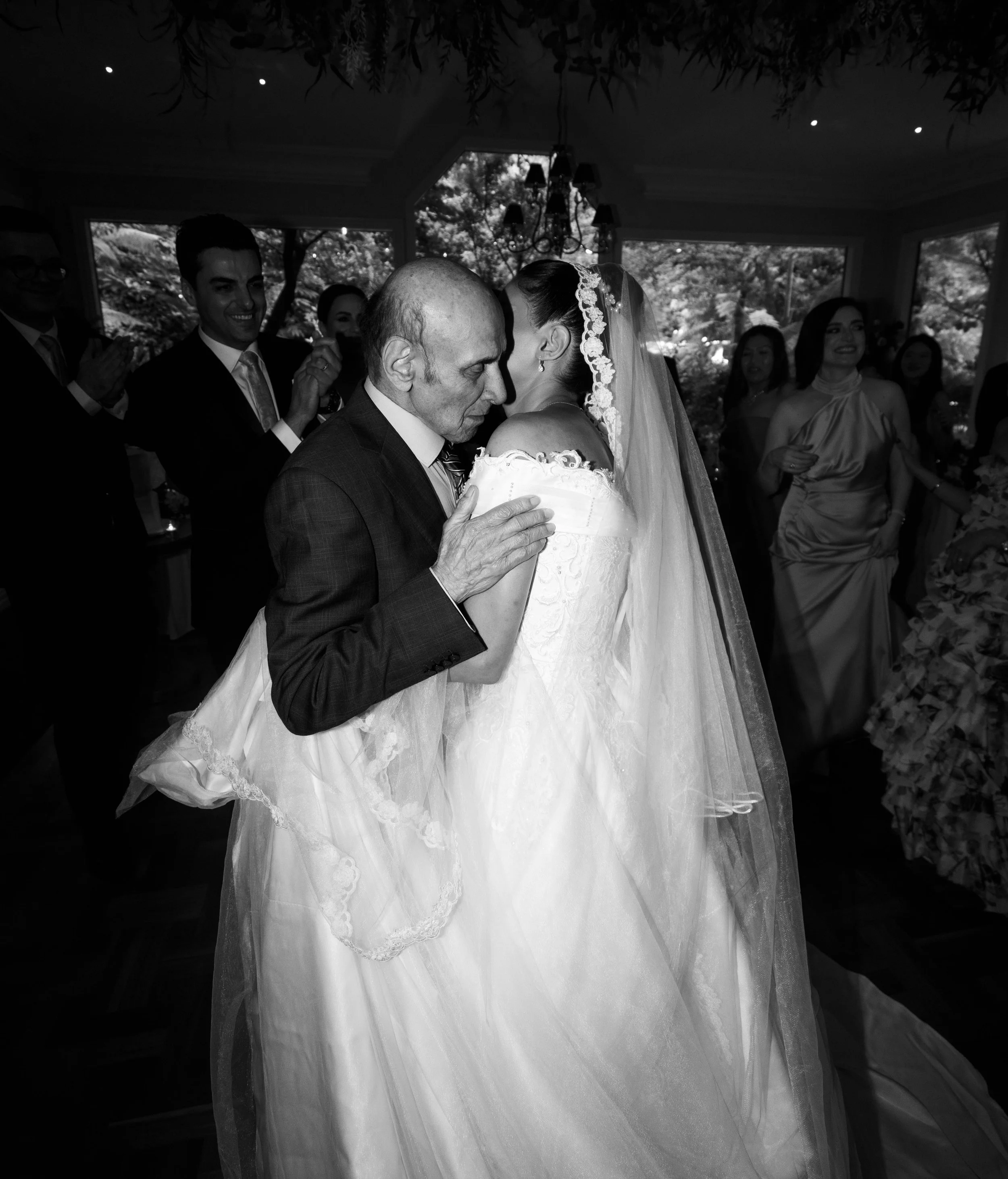 A black and white photo of a bride and an older man, possibly her father, sharing a dance at a wedding reception. The bride is wearing a wedding gown with lace details and a veil, while the older man is dressed in a suit. Several guests are watching 