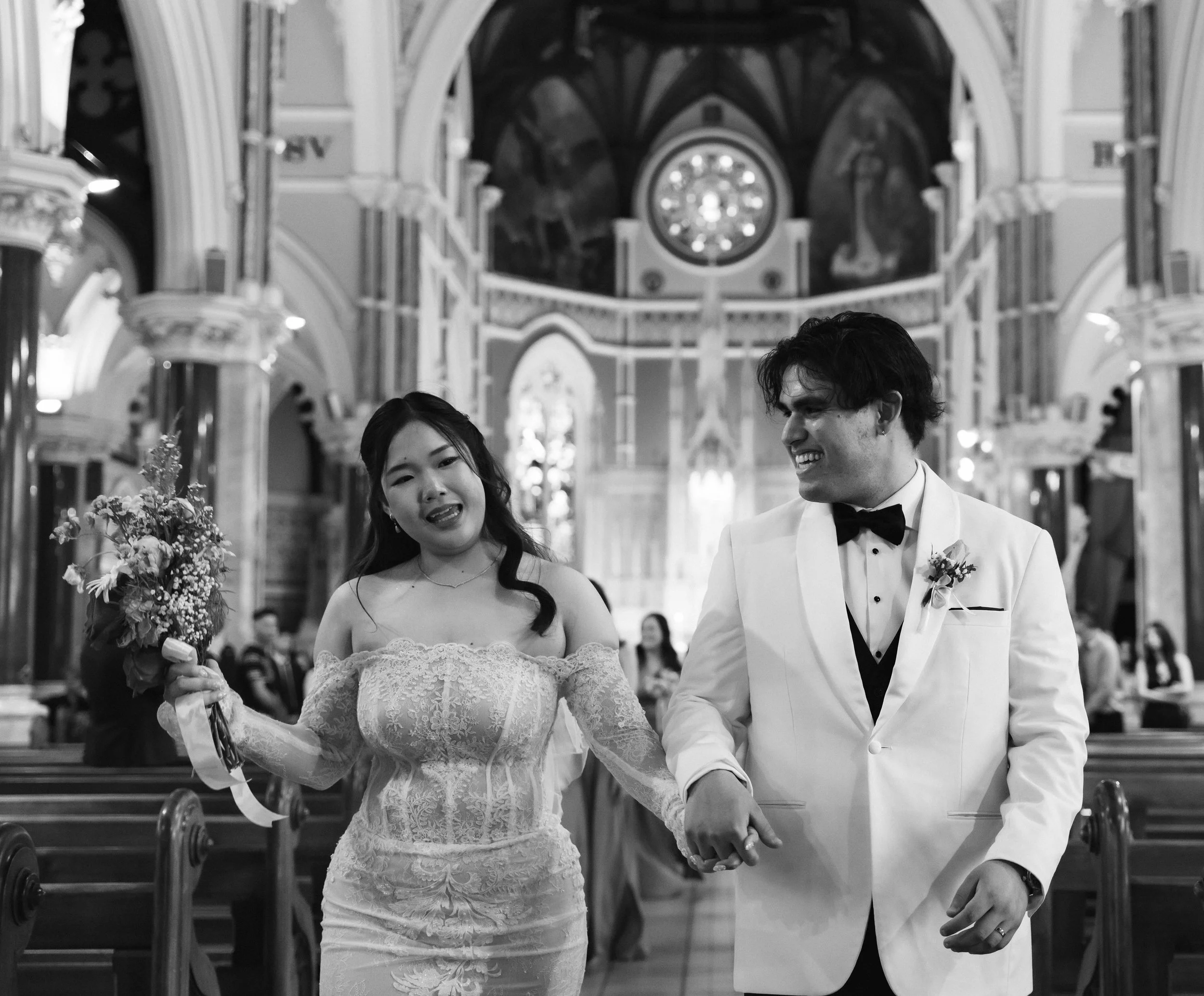 A black and white photo of a bride and groom holding hands during a wedding ceremony inside a church.