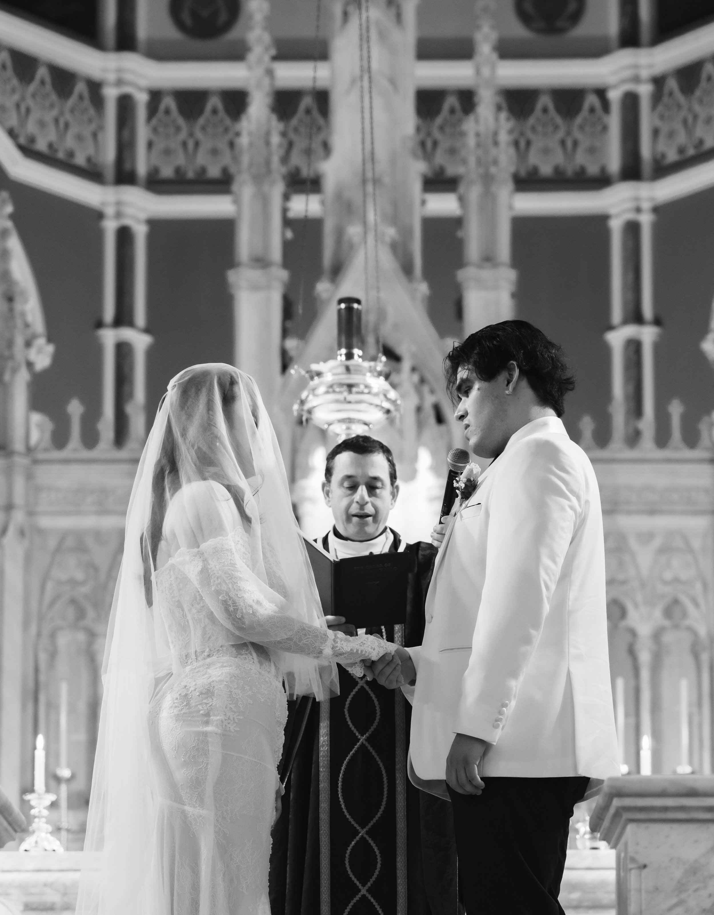 A black and white photo of a wedding ceremony taking place in a church, with a bride and groom standing at the altar facing each other, an officiant, and ornate architectural details in the background.