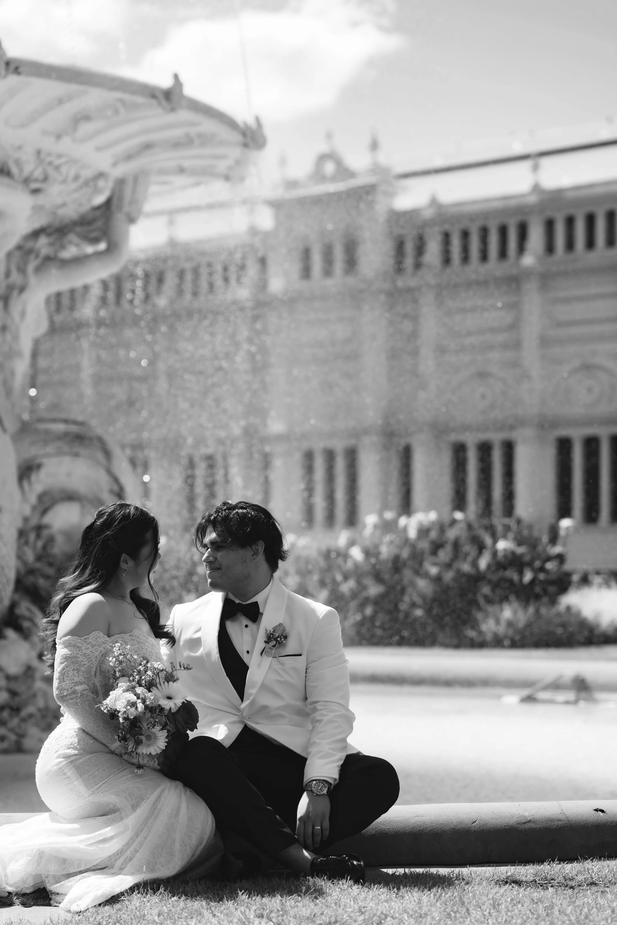 A bride and groom sitting together outside in front of a historic building, with the bride holding a bouquet and both dressed in wedding attire.
