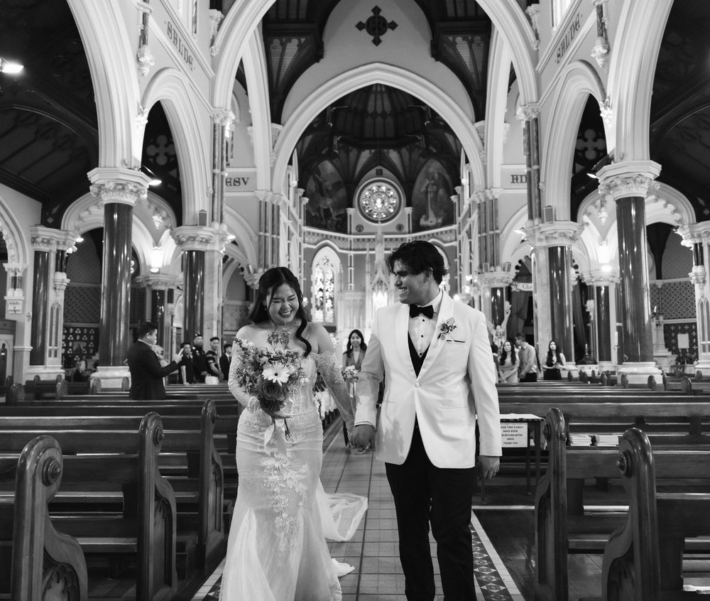 A black and white photo of a bride and groom holding hands inside a church with high vaulted ceilings and stained glass windows.