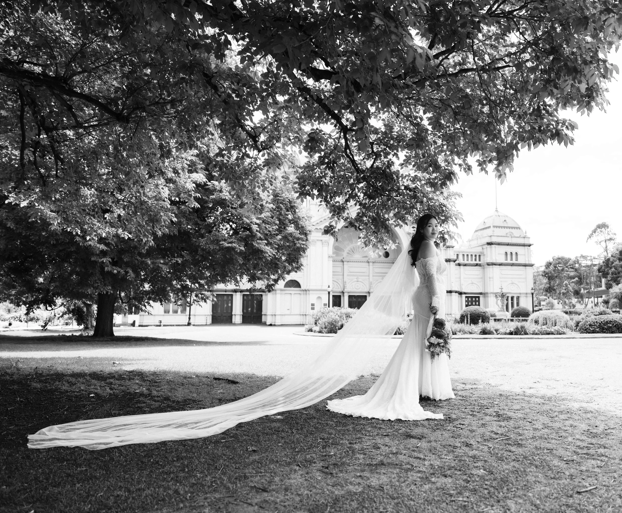A bride in a wedding gown standing outdoors under a large tree, holding a bouquet, with a historic building in the background in black and white.
