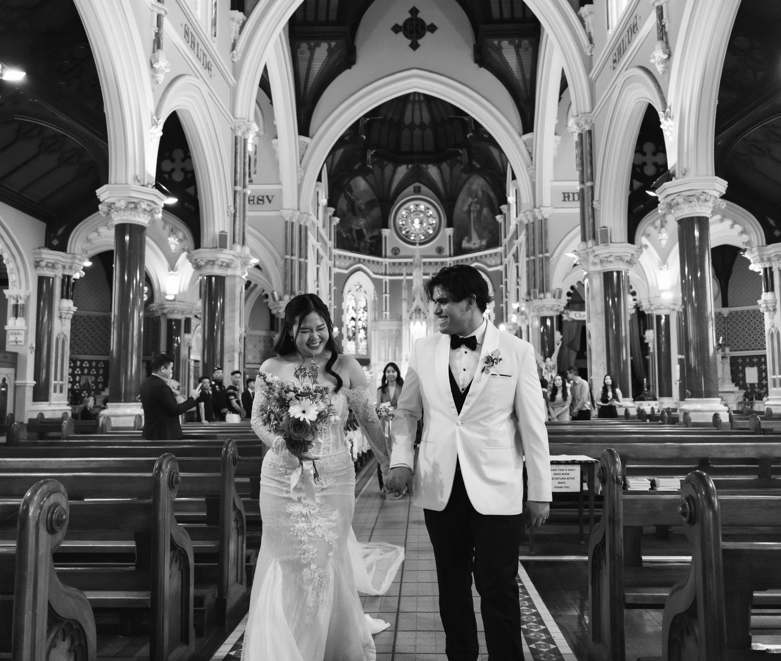A bride and groom holding hands and smiling inside a church with high vaulted ceilings, ornate arches, and stained glass windows.