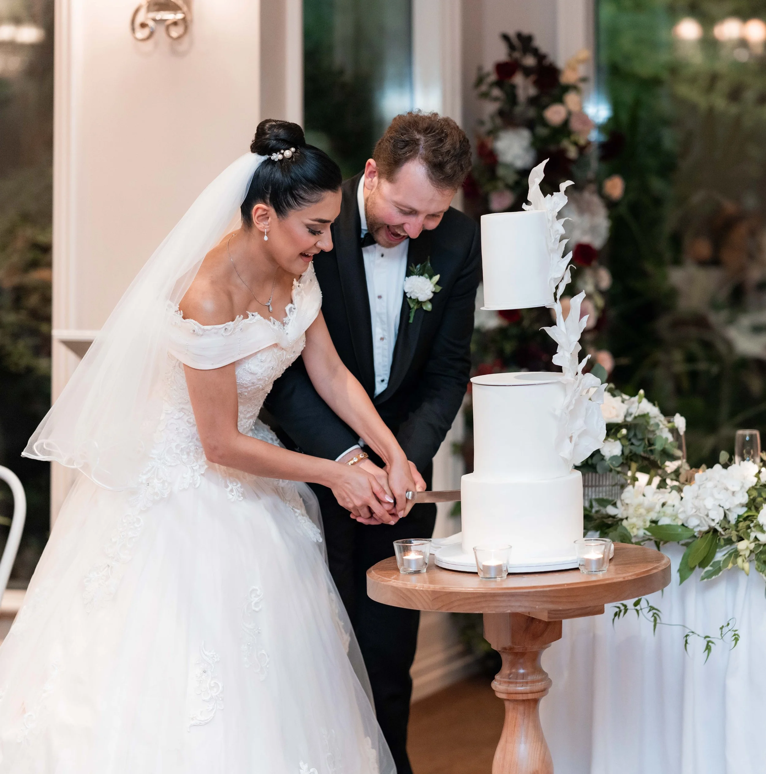 A bride and groom at their wedding ceremony, holding hands and cutting a white wedding cake with white flowers on it, standing beside a wooden table with candles, in a decorated indoor venue with floral arrangements.