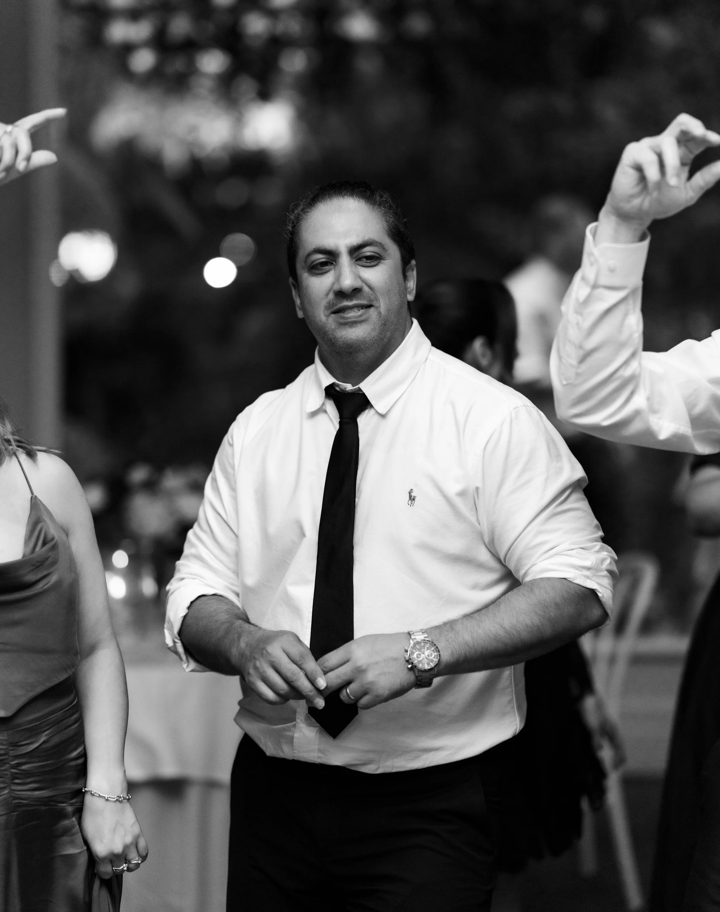 A black-and-white photo of a man in a white dress shirt and black tie at a social event. He is looking at the camera with a slight smile, wearing a watch on his left wrist. Other people and blurred lights are visible in the background.