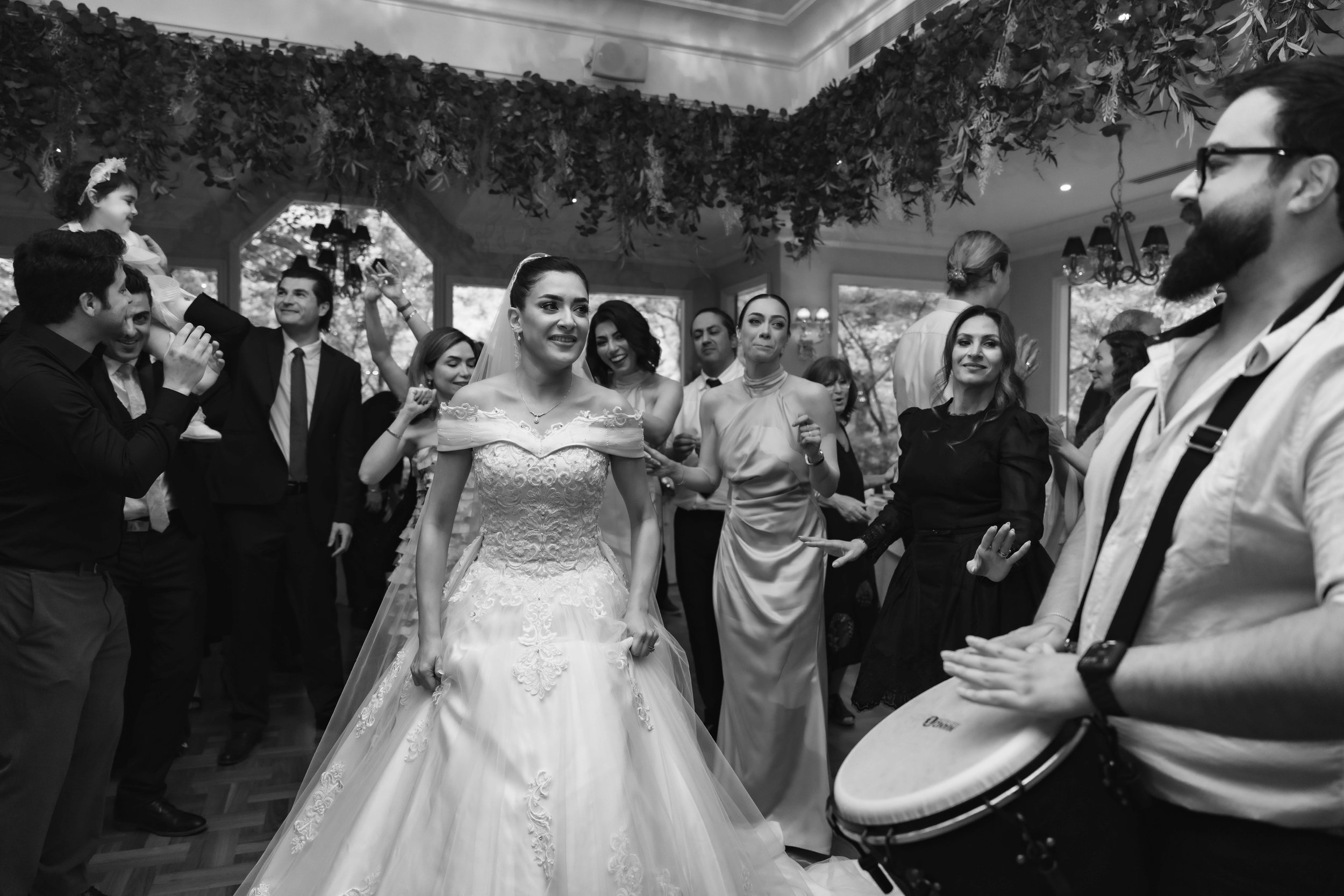Black and white photo of a wedding reception with a bride dancing in a wedding gown, surrounded by guests. Some guests are smiling and talking, with woman in a sleeveless dress near the bride, and a man playing a guitar on the right side of the image