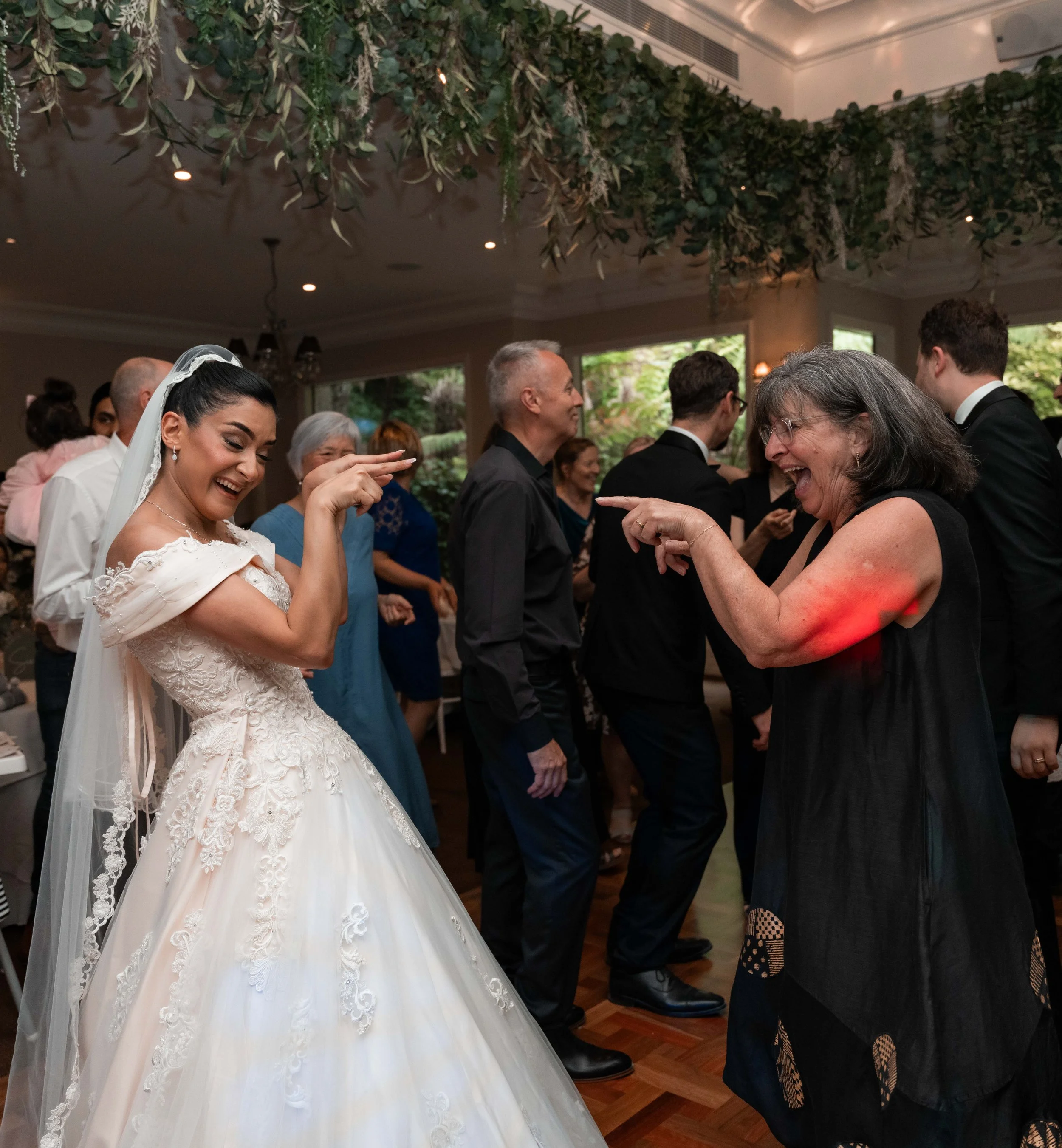People dancing at a wedding reception indoors, with a bedroom dress in the foreground and a ceiling decorated with green leaves and white flowers.
