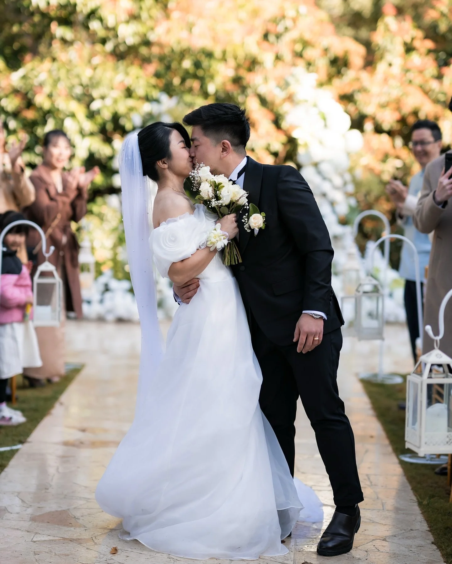 A bride and groom kissing during their outdoor wedding ceremony, with guests smiling and clapping in the background.