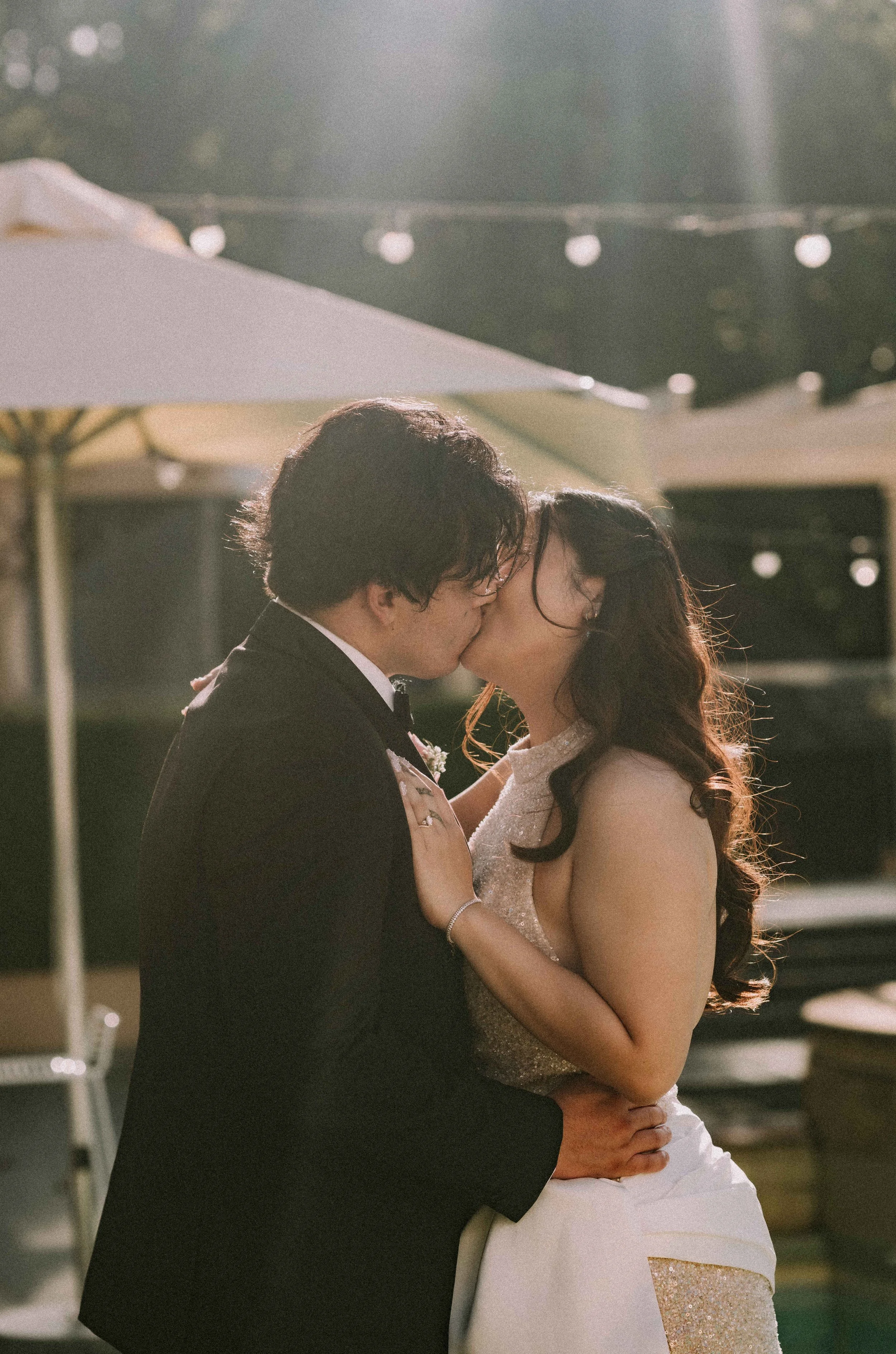 A bride and groom kissing at an outdoor wedding reception, with umbrellas and string lights visible in the background.
