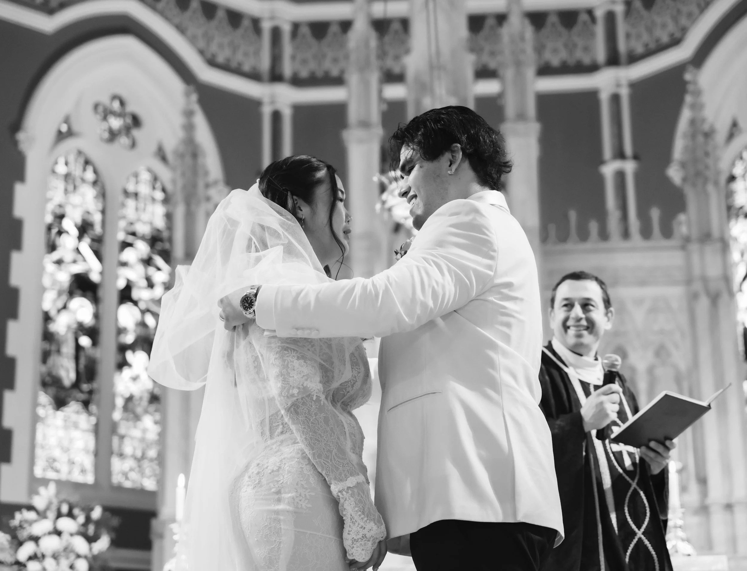 A bride and groom are smiling and embracing during their wedding ceremony inside a church with stained glass windows and ornate architecture.