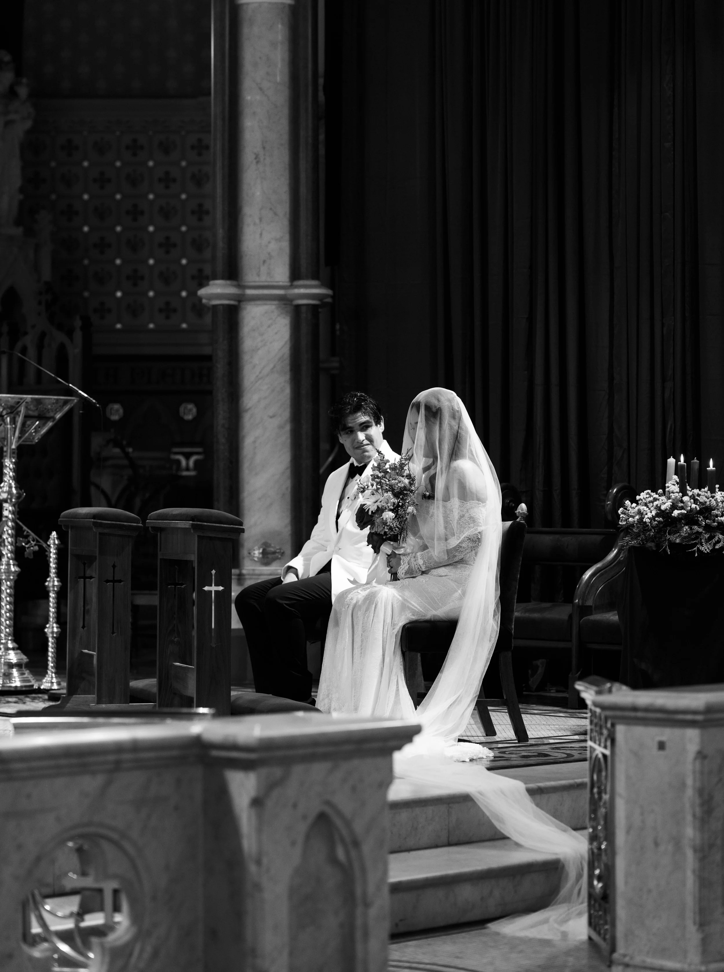 A black and white photo of a bride and groom sitting in a church during their wedding ceremony. The bride is wearing a wedding dress and veil, holding a bouquet, and the groom is wearing a tuxedo. They are seated on a bench with religious candles and