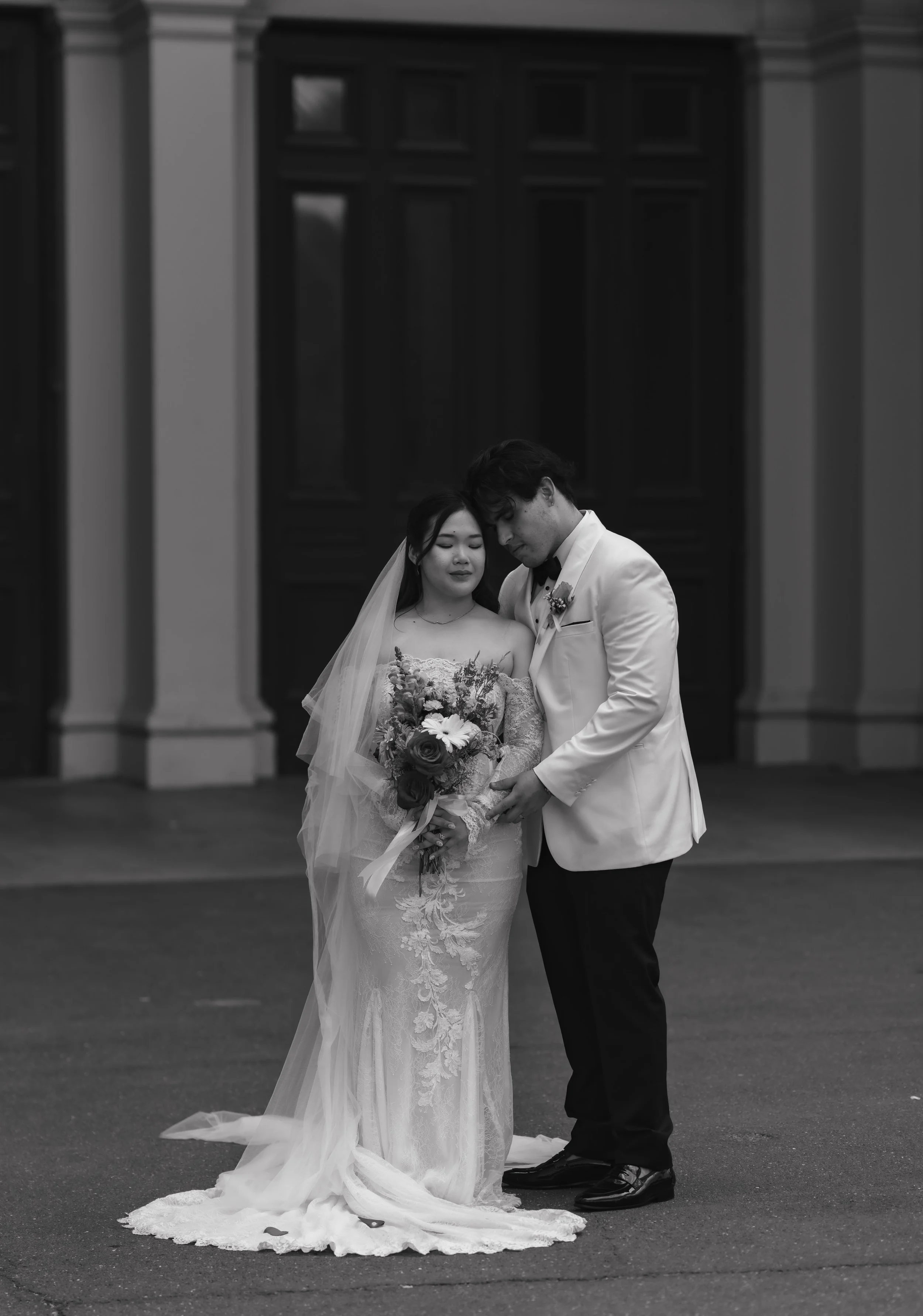 A bride and groom standing close together on a street, eyes closed, with heads touching, in front of a building with large dark wooden doors. The bride is holding a bouquet, wearing a lace wedding dress and veil, while the groom is in a white tuxedo 