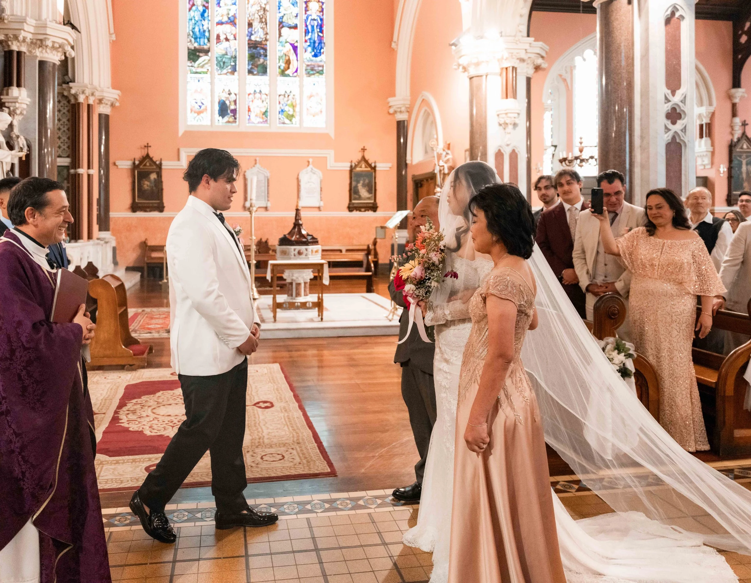 A wedding ceremony inside a church with stained glass windows, featuring a bride in a white gown and veil, and a groom in a white tuxedo with a black tie, facing each other. Guests, including a woman in a peach dress and a man in a beige suit, are wa