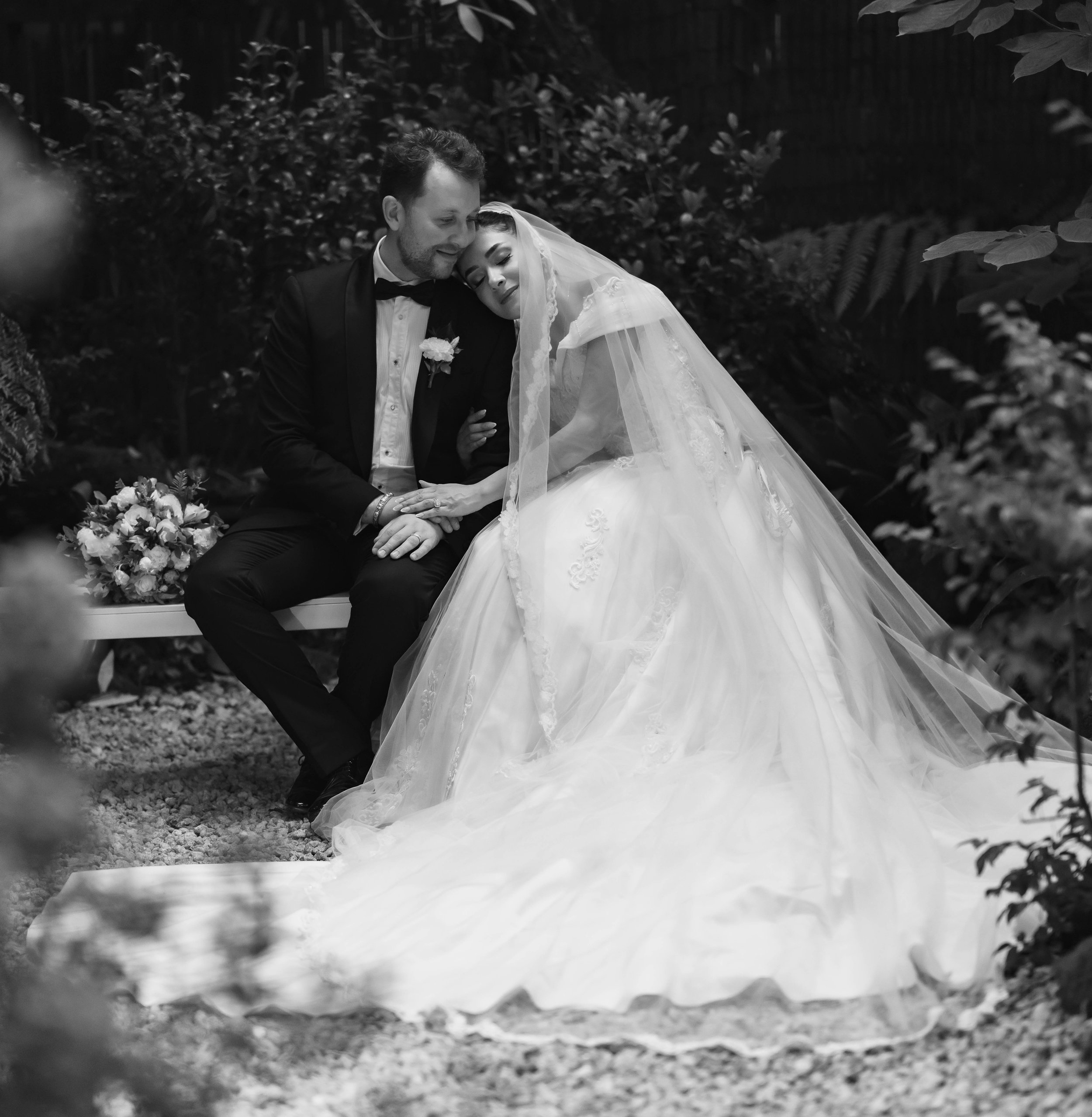 Black and white photo of a bride and groom sitting close together outdoors, with the bride resting her head on the groom's shoulder. The groom is wearing a tuxedo with a bow tie, and the bride is in a wedding dress with a veil. They are surrounded by