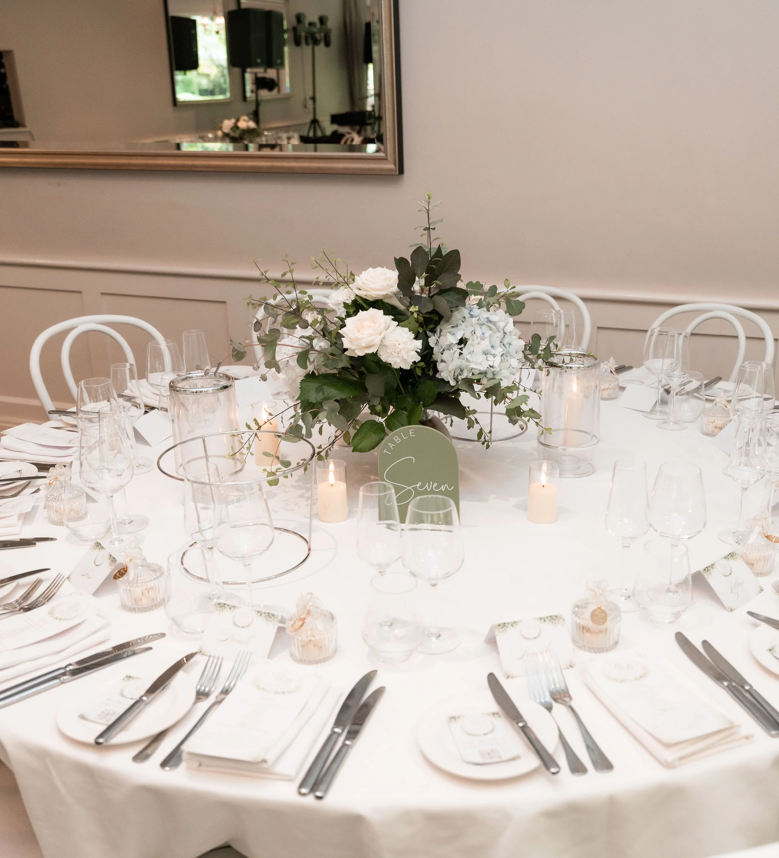 A round white dining table decorated with a floral centerpiece featuring white roses, hydrangeas, and greenery. The table is set with white plates, silverware, wine glasses, and tealight candles in glass holders. A labeled place card reads 'Table Sev