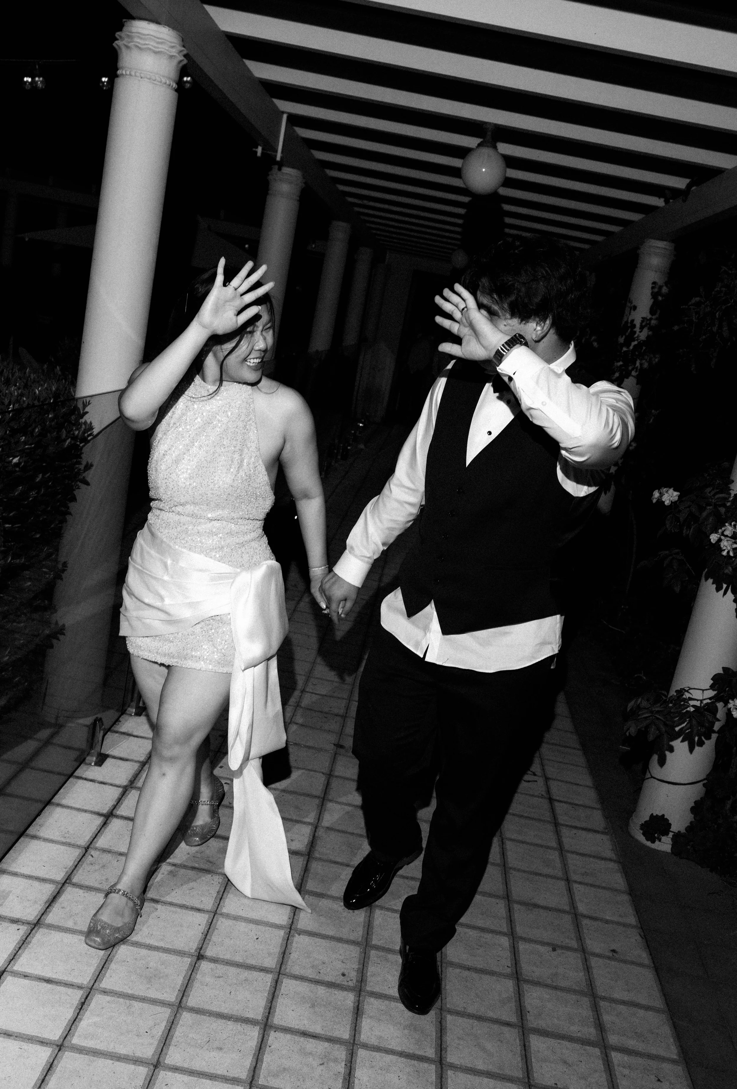 A black and white photo of a woman in a dress and a man in a vest and shirt holding hands on a tiled porch, smiling and raising their other hands as if greeting or waving.