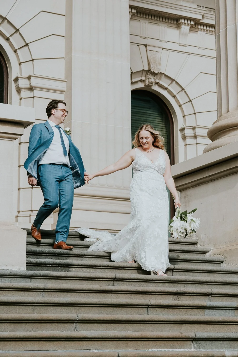 A bride and groom walking down the steps of a grand building, holding hands.