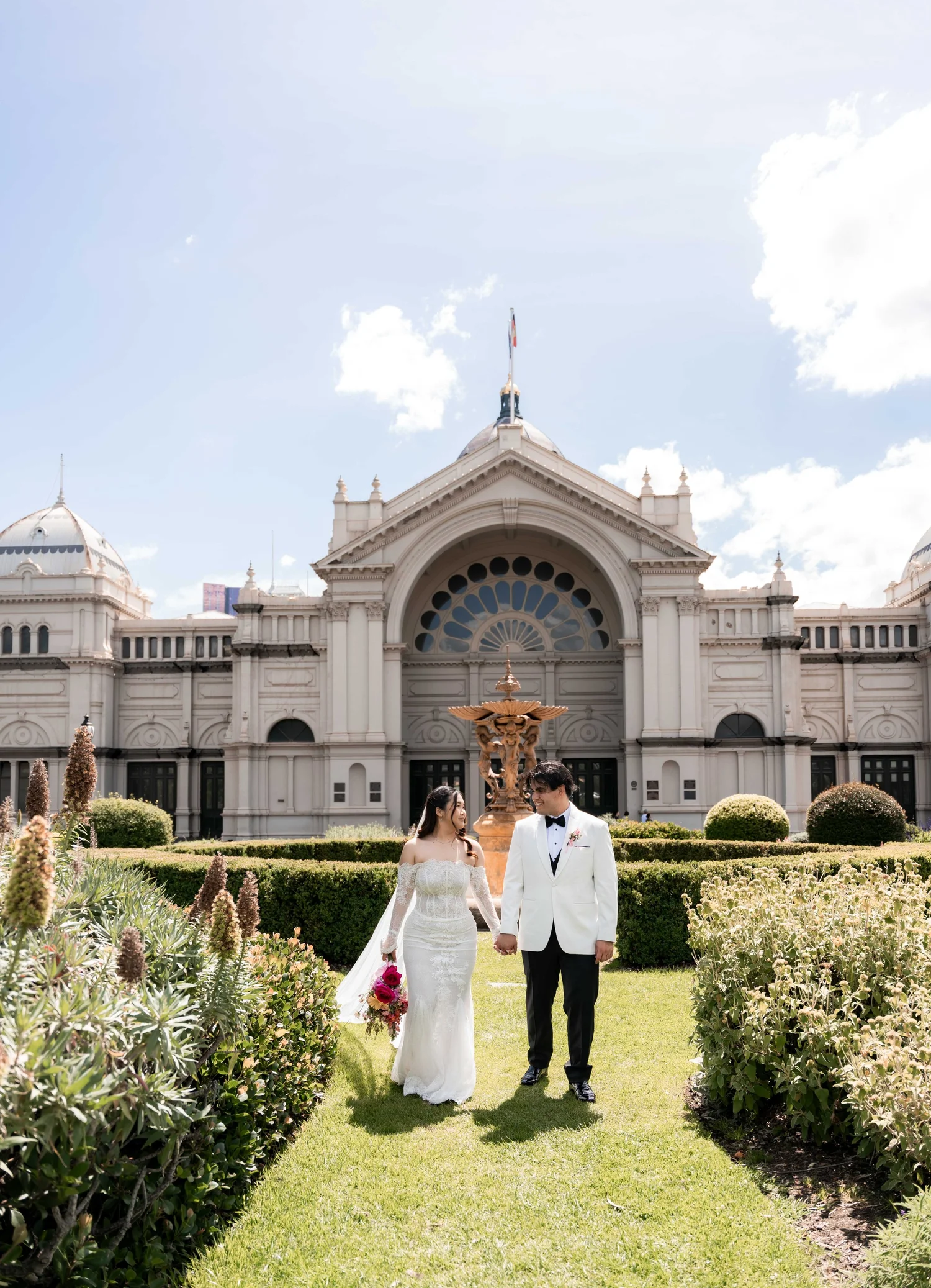 A bride and groom walking hand in hand through a garden in front of a grand historic building.