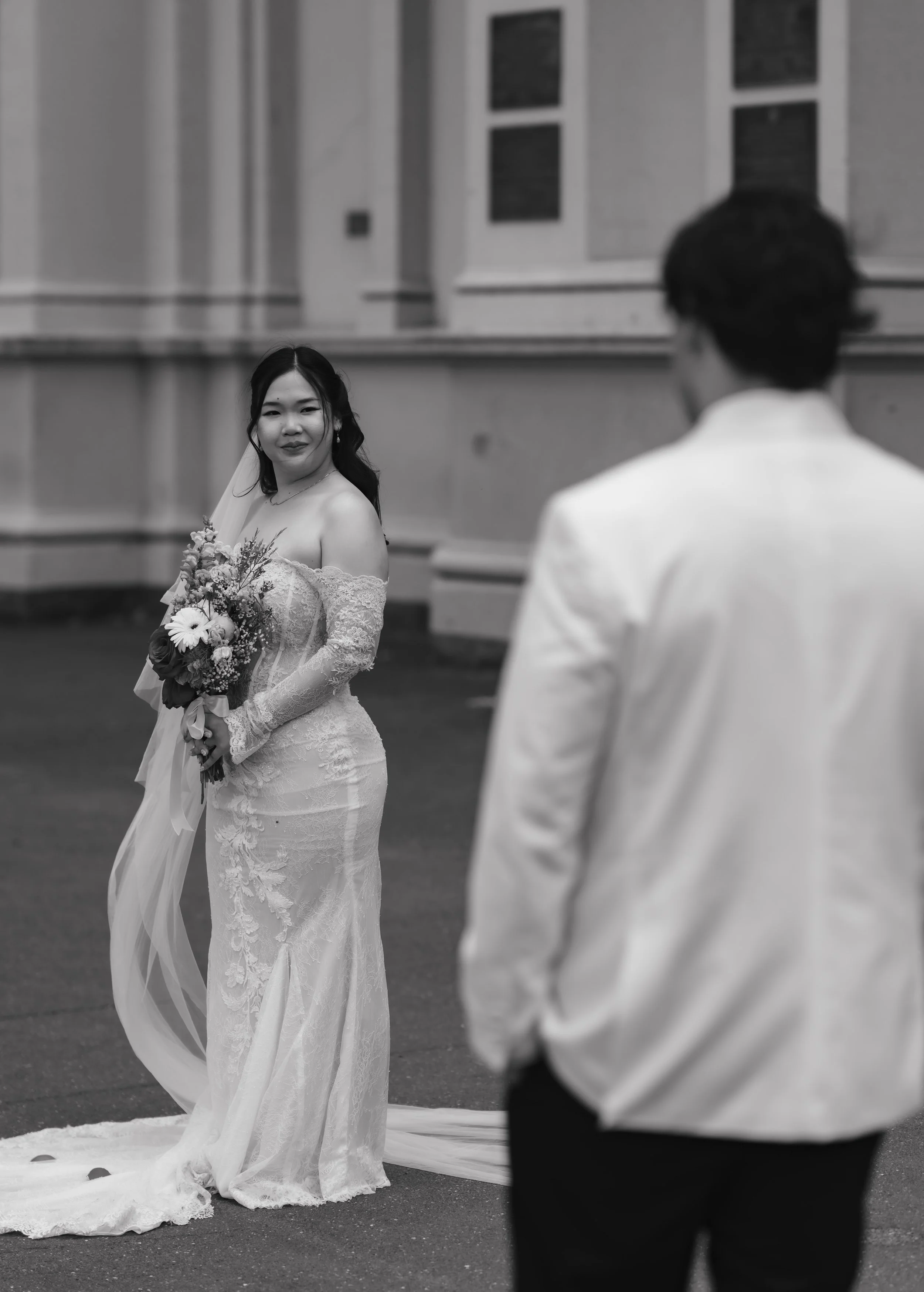 Black and white photo of a bride in a lace wedding dress holding a bouquet, smiling at a man in a white tuxedo, outside near a building with windows.