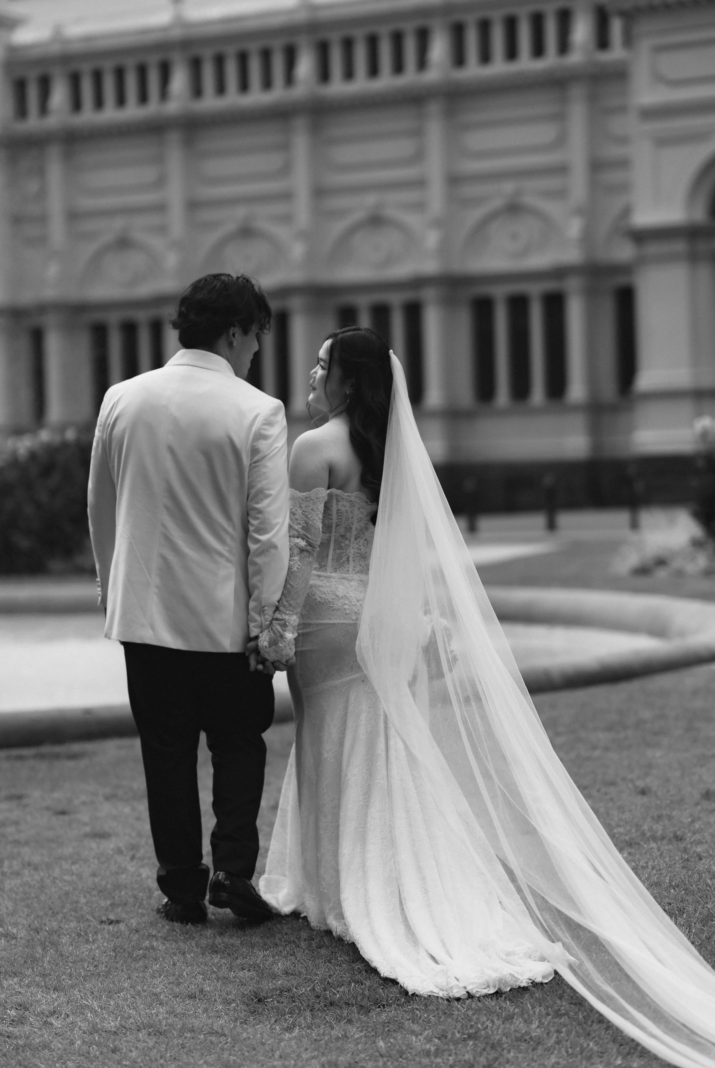 A black and white photo of a bride and groom holding hands outdoors with a historic building in the background.