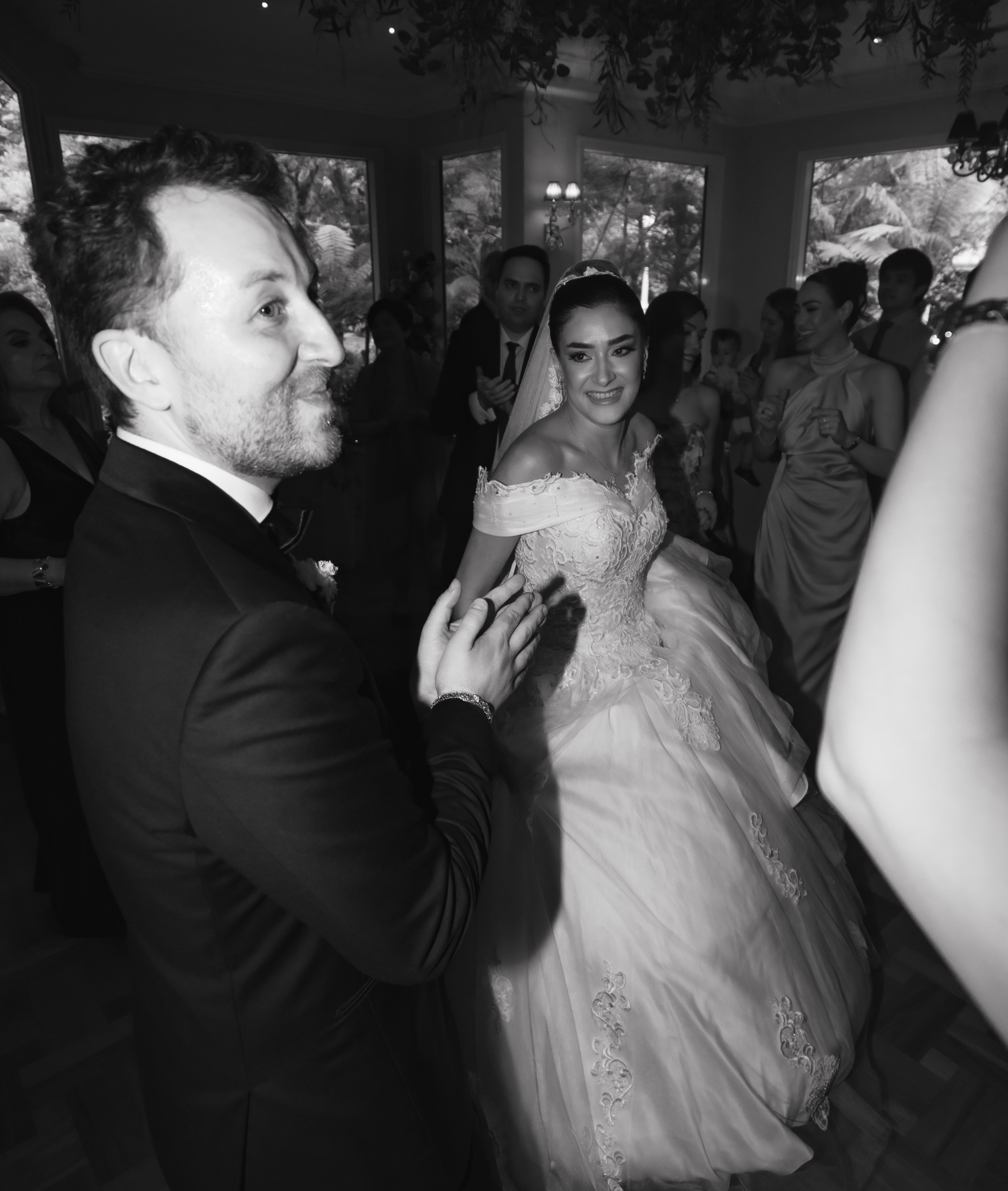 Black and white photo of a wedding celebration with a bride in a wedding dress sitting, surrounded by guests, some smiling and watching.