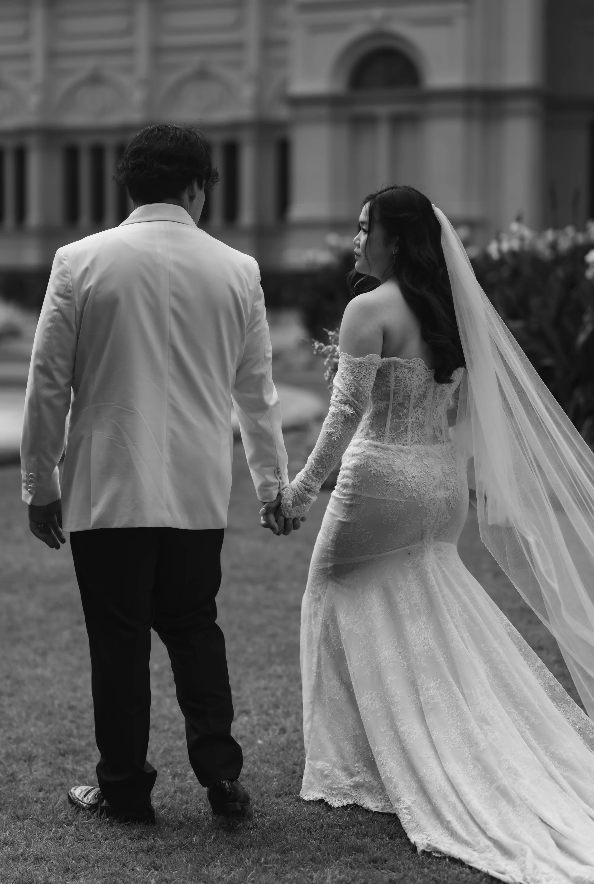 A bride in a lace wedding gown with a veil holding hands with a groom in a white suit jacket, standing outdoors in front of a building.