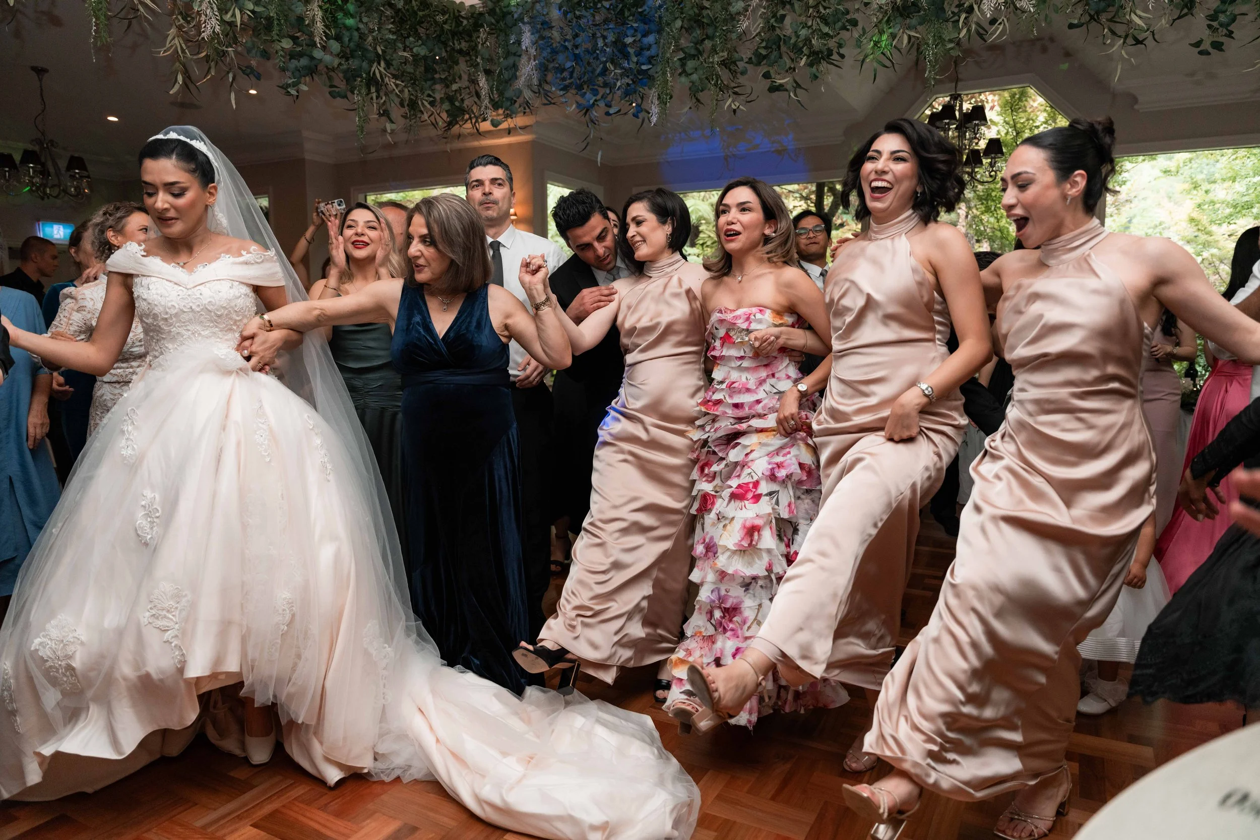 A group of people at a wedding reception dancing and celebrating indoors with big windows showing greenery outside.