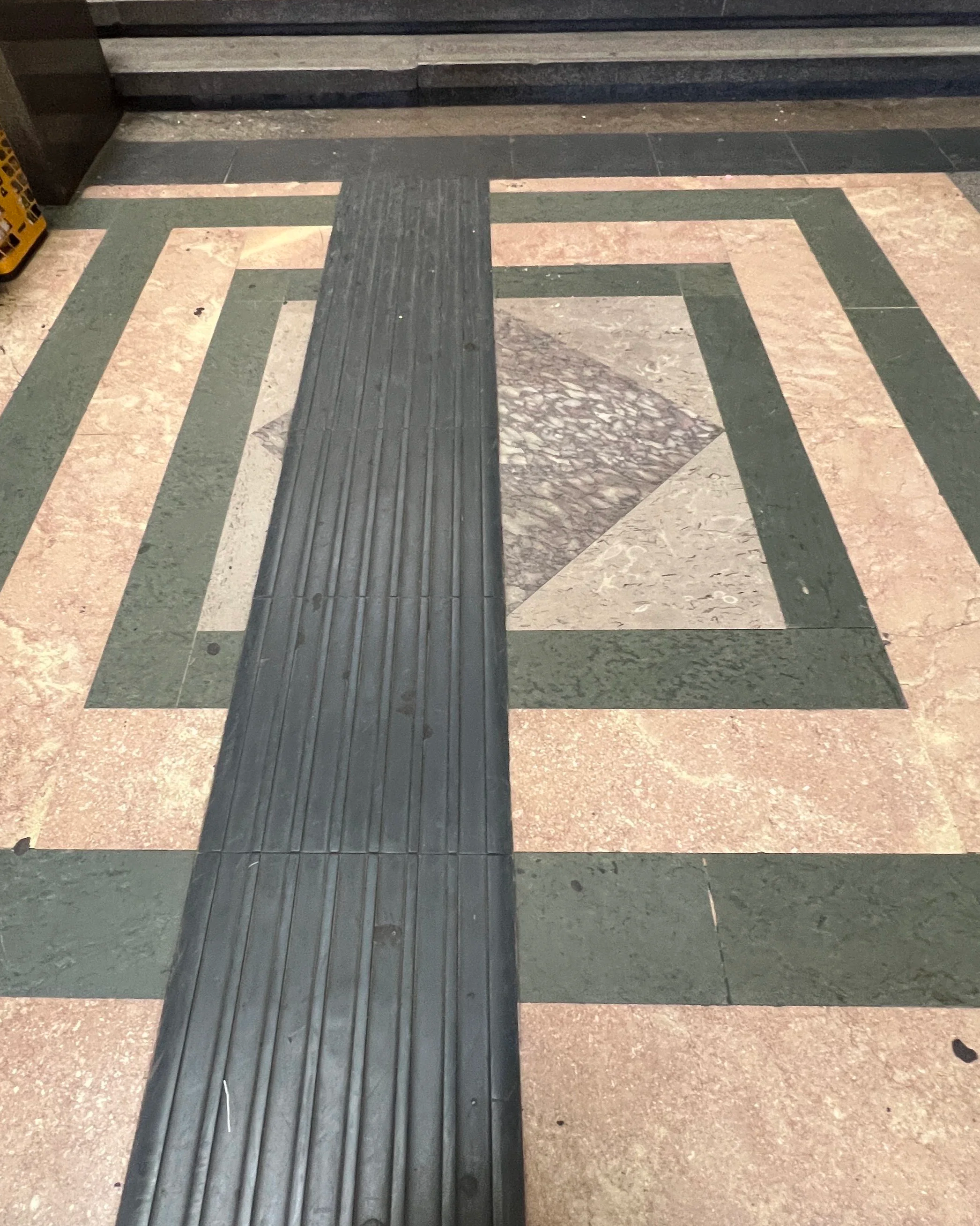 A tiled floor with a black tactile paving strip and geometric patterns in various shades of beige, cream, and green surrounding it.