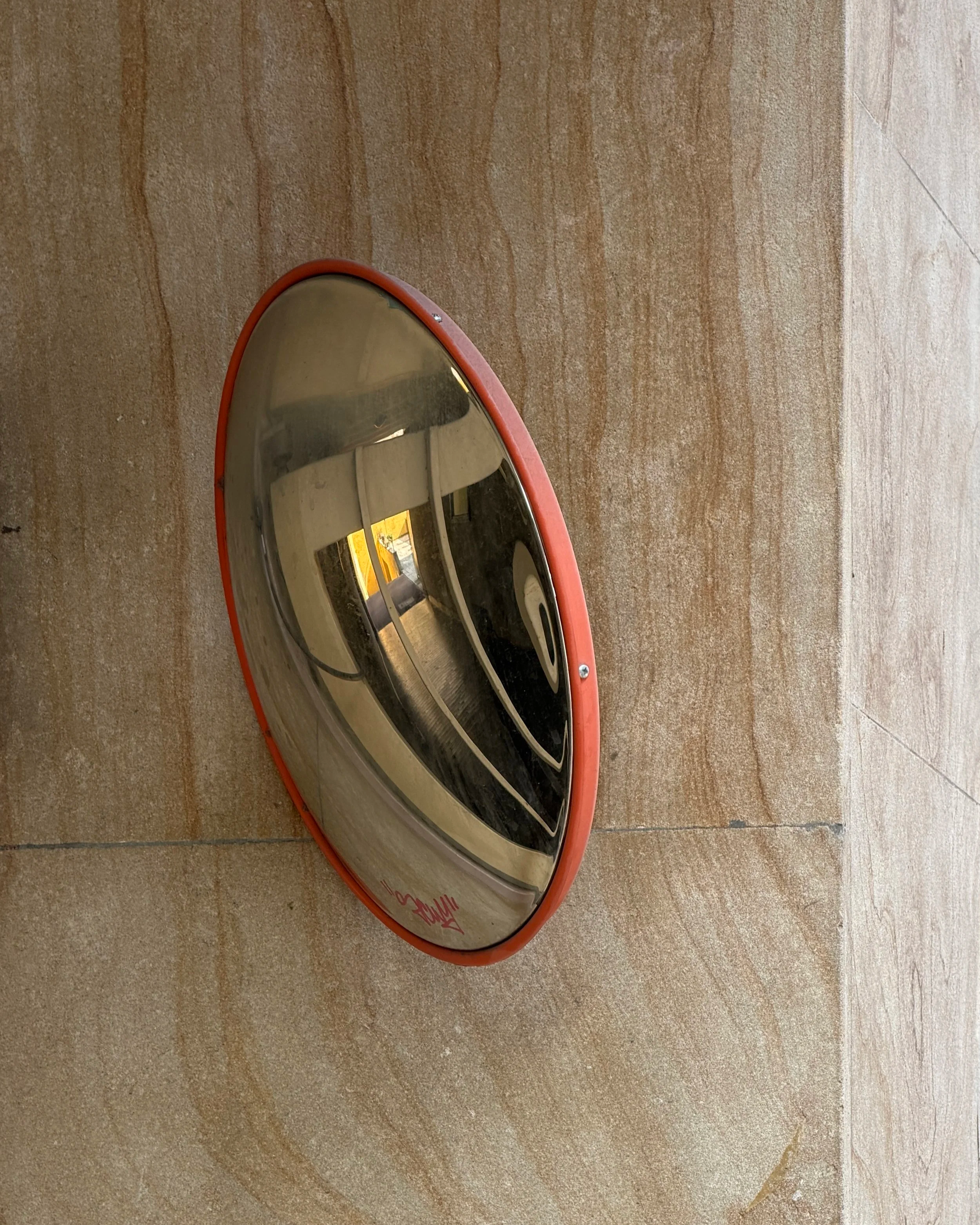 A close-up of a circular mirror with a red frame, lying on a beige wooden surface. The mirror's reflection shows a room with beige walls, ceiling, and fluorescent lighting.