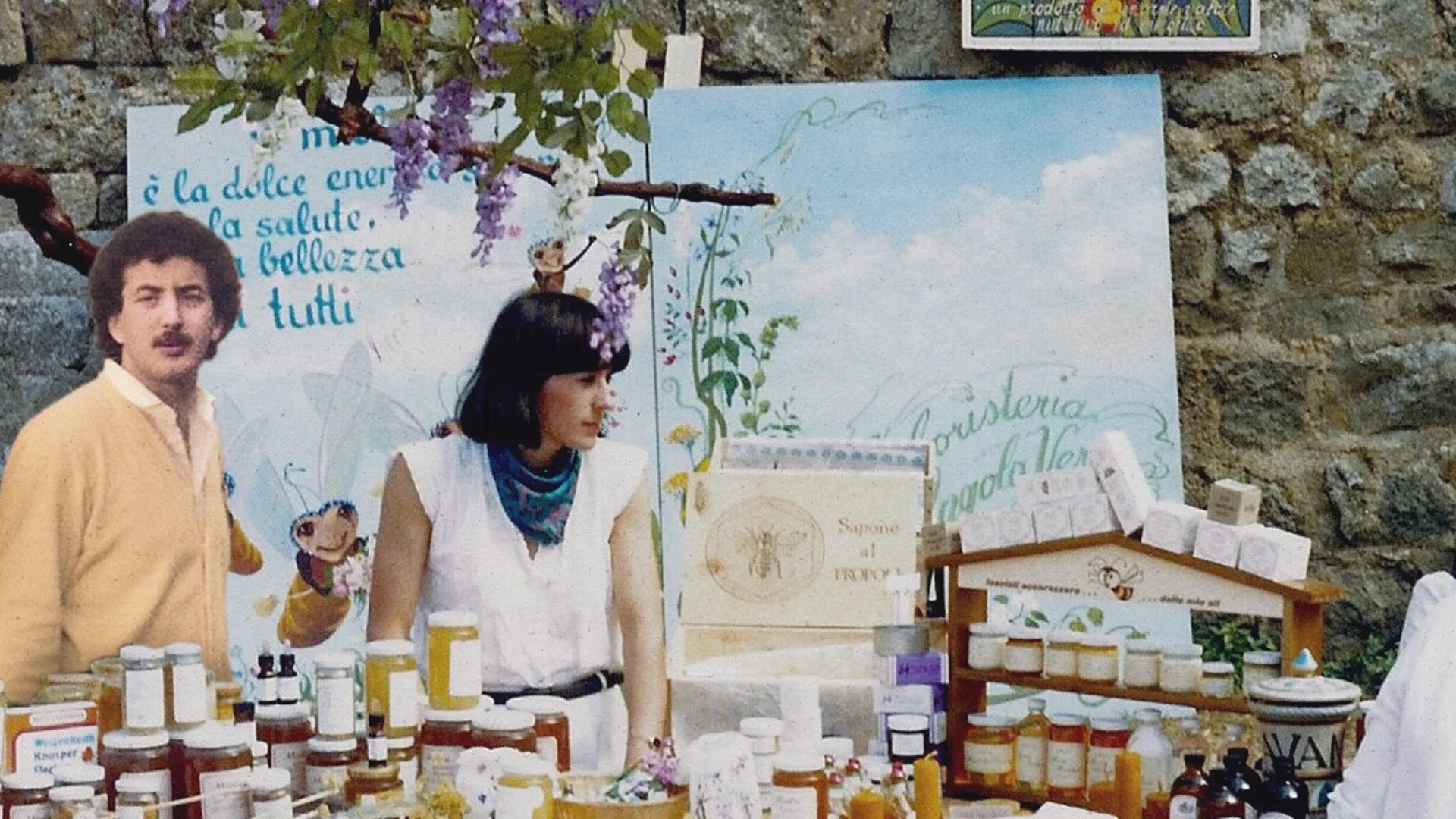 Two women and a man at a stall selling honey and herbal products, with a stone wall and decorative signs in the background.