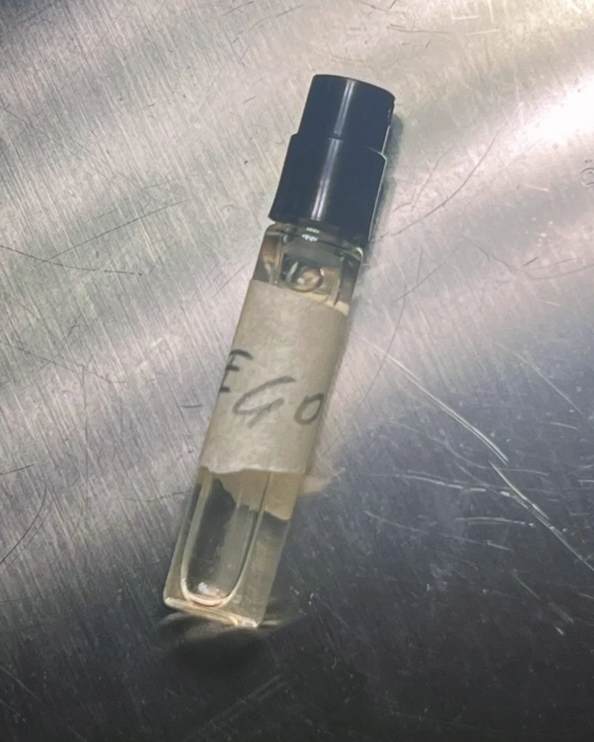 A small vial or test tube with a black cap, sitting on a metallic surface. The vial has a handwritten label that appears to say 'EGO'.