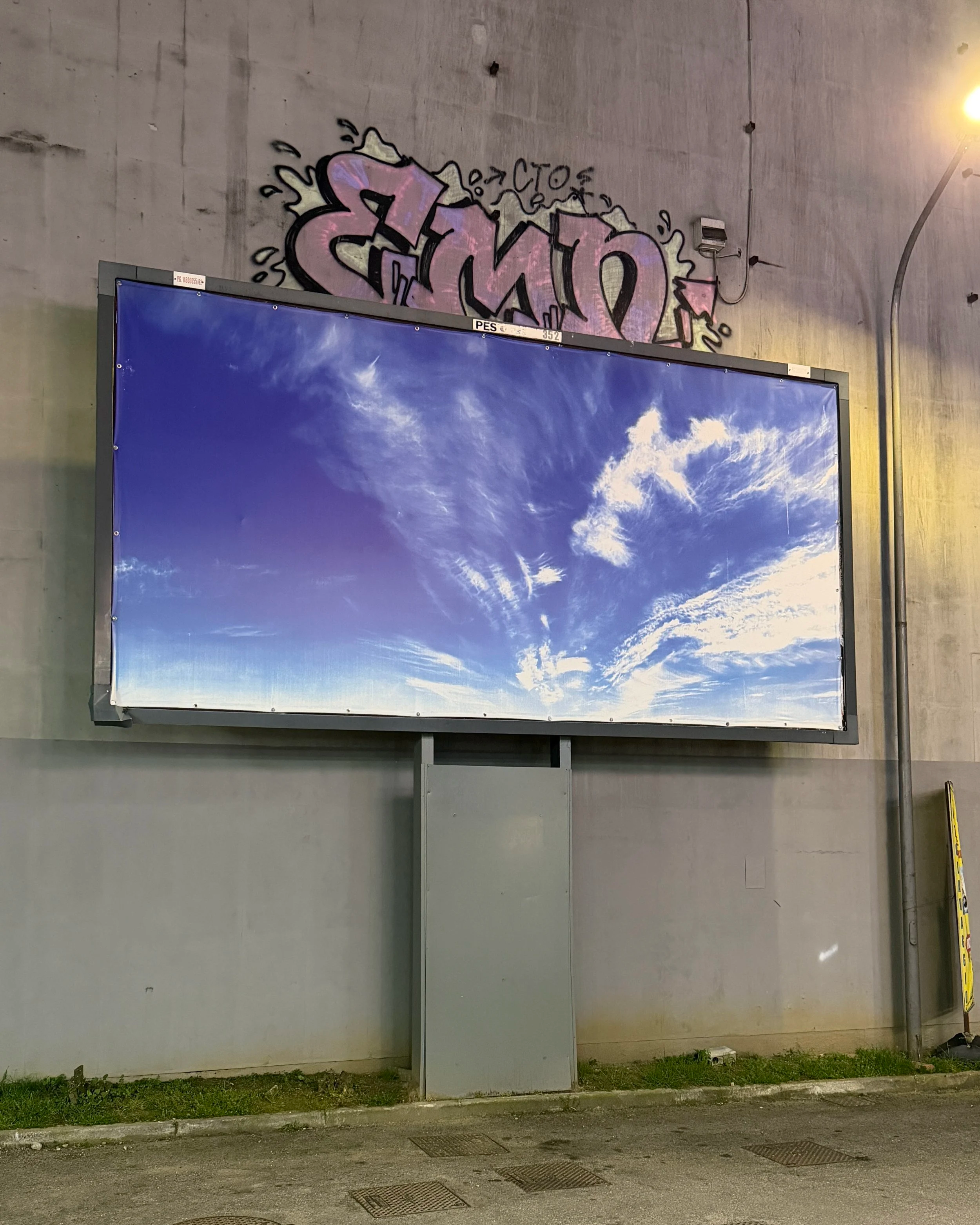 Graffiti art on a concrete wall with colorful lettering and arrow pointing to the right, adjacent to a billboard displaying a sky with clouds.