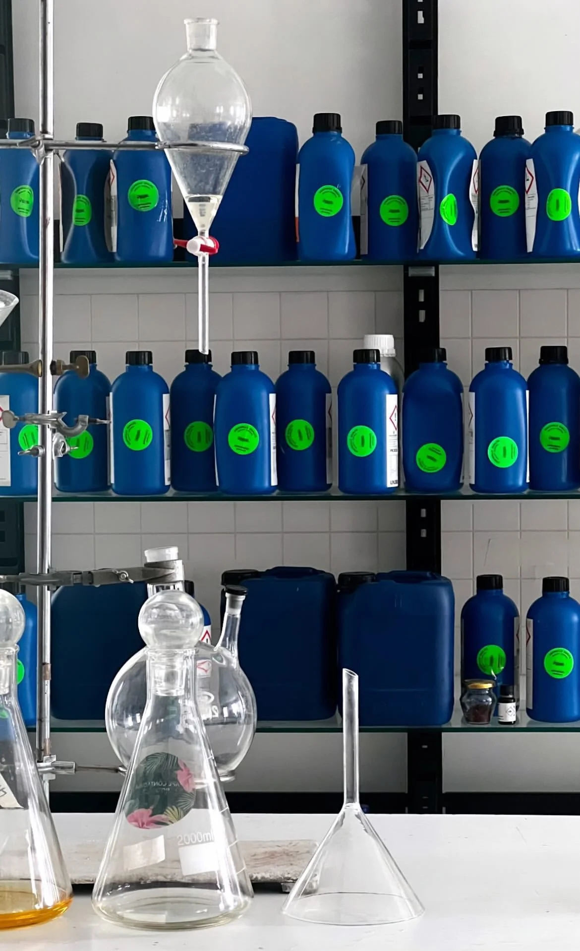 Lab shelves with blue chemical bottles labeled with green safety labels, and glass laboratory equipment including flasks and beakers on a white work surface.
