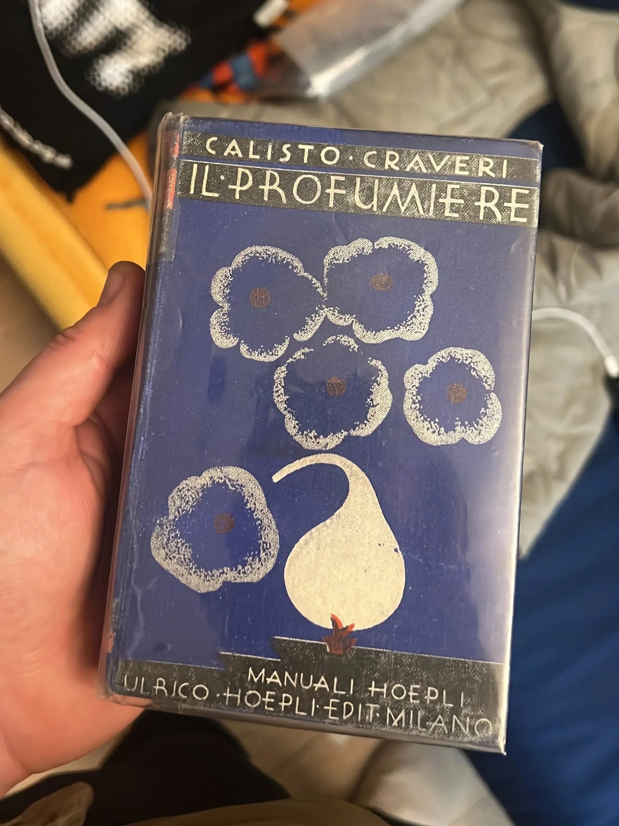 A person holding a black and blue box with white embroidered flowers and gold lettering. The box features the names Calisto Craveri and Il Profumo Re, with additional Spanish and Italian text at the bottom.