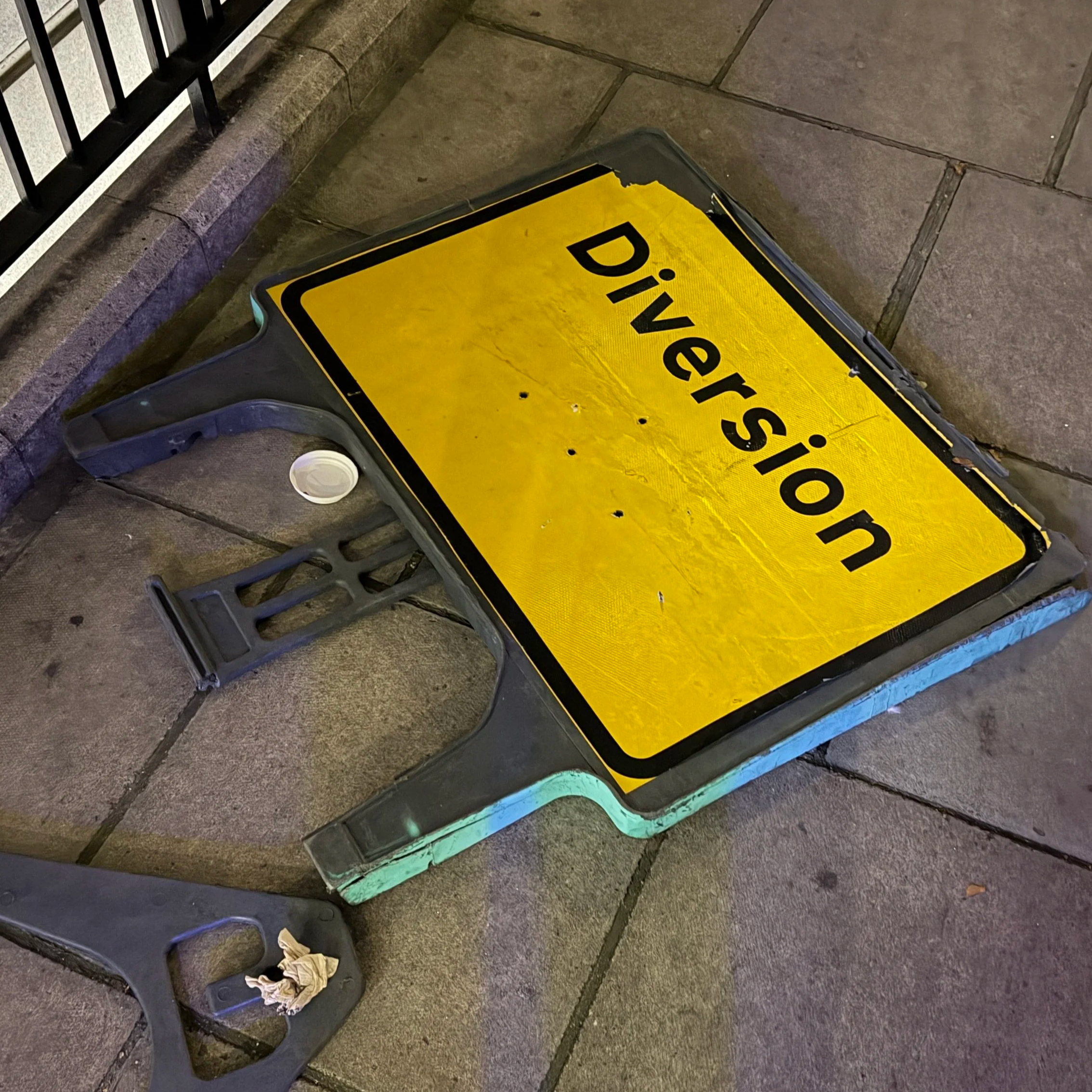 A yellow and black caution sign fallen on a brick sidewalk. The sign reads ' Dividend'. There is a trash bin with some paper and a cup next to the sign. The sidewalk is made of rectangular stone tiles, and there is a black metal fence in the background.