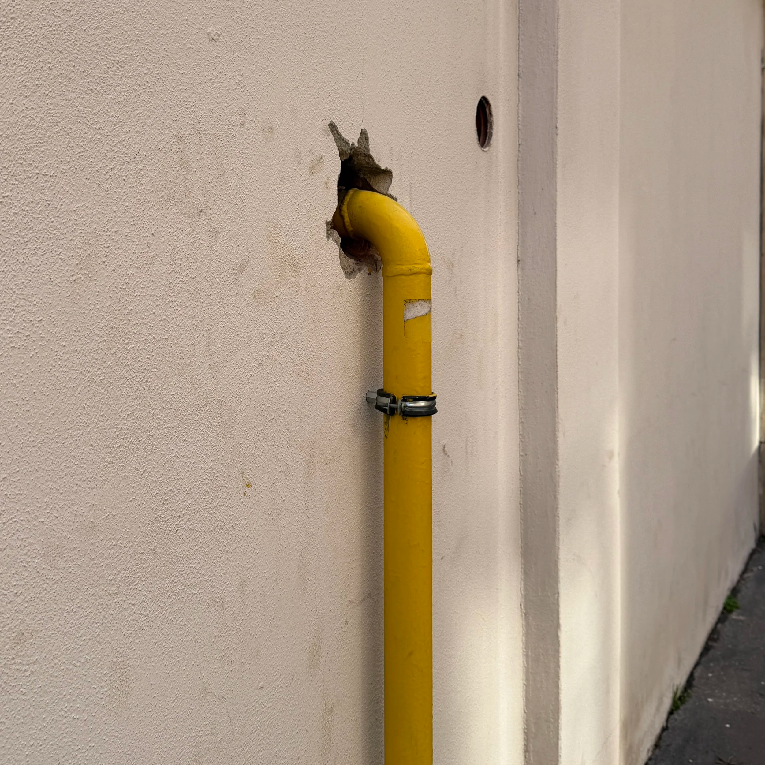 A yellow gas pipe protrudes from a beige textured wall, with an opening in the wall nearby. The scene is outdoors with a sidewalk and some greenery visible at the bottom right corner.
