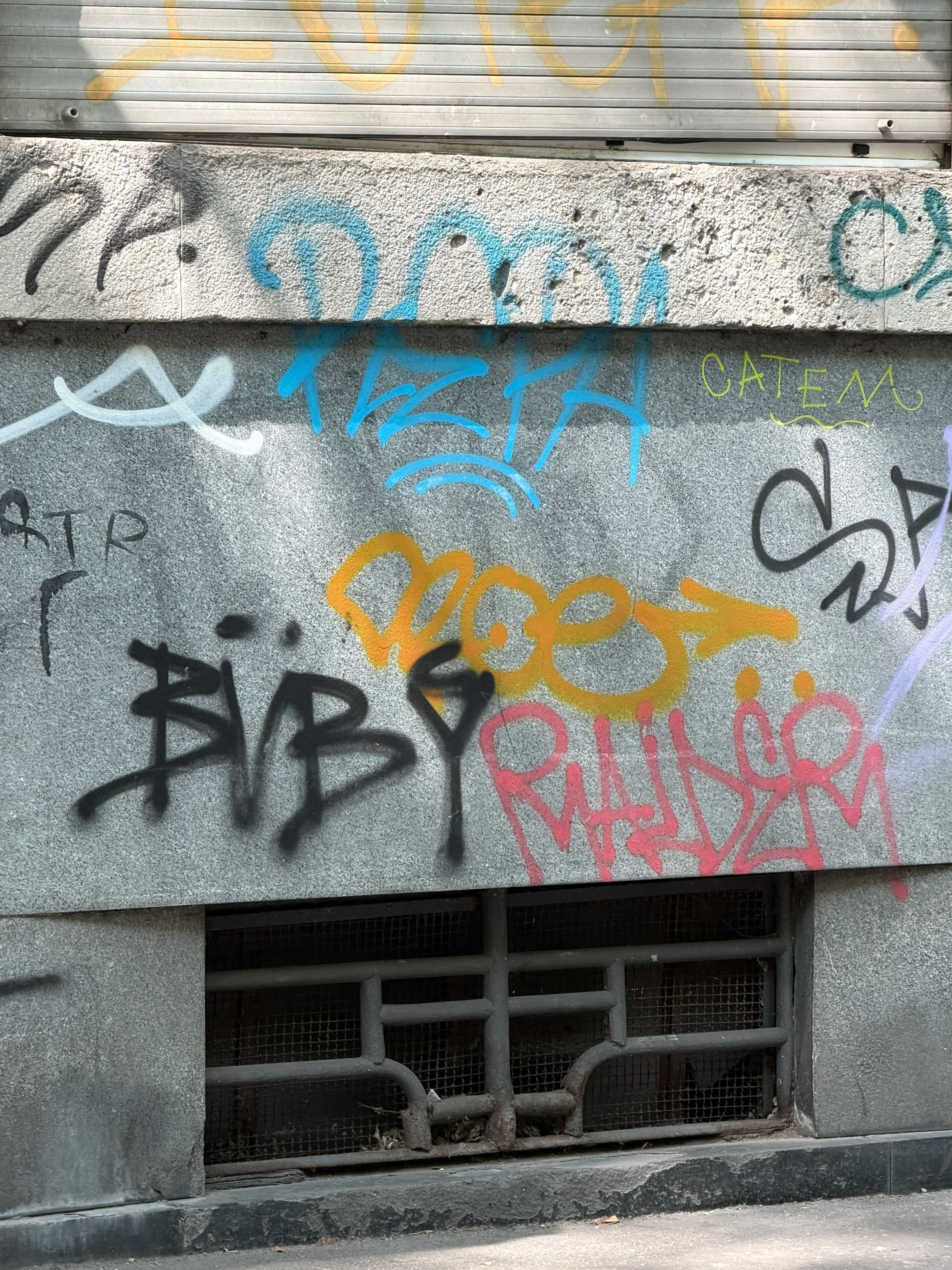 Street-level view of a metal grate and concrete structure with colorful graffiti tags painted on the wall above and around it.