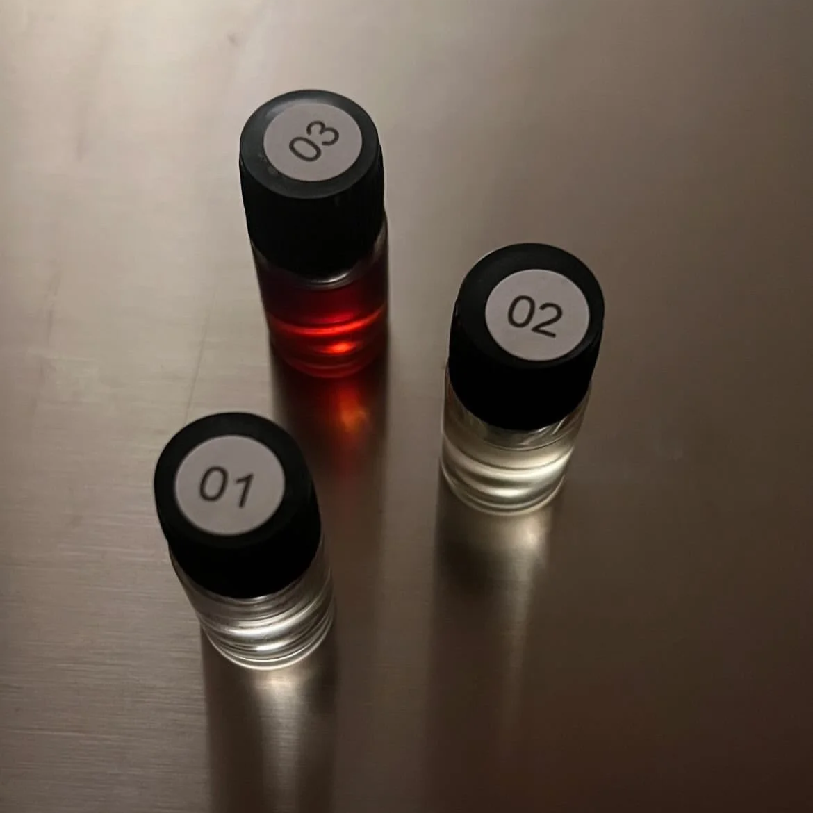 Three small bottles with numbered black caps labeled 01, 02, and 03, containing clear, yellow, and red liquids, respectively, placed on a metallic surface.