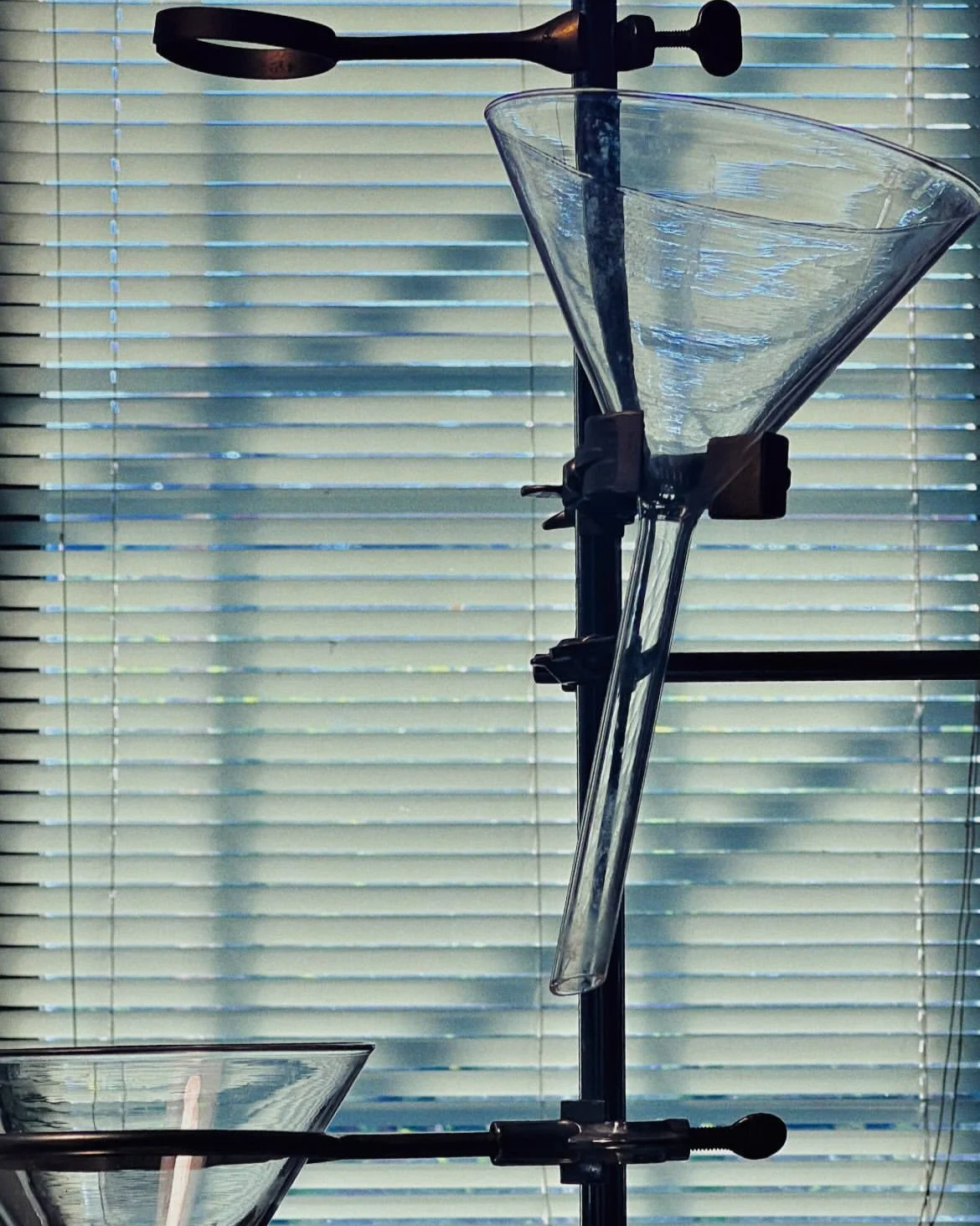 Laboratory equipment for distillation or chromatography, including a glass funnel, a glass container, and stand, in front of a window with blinds.