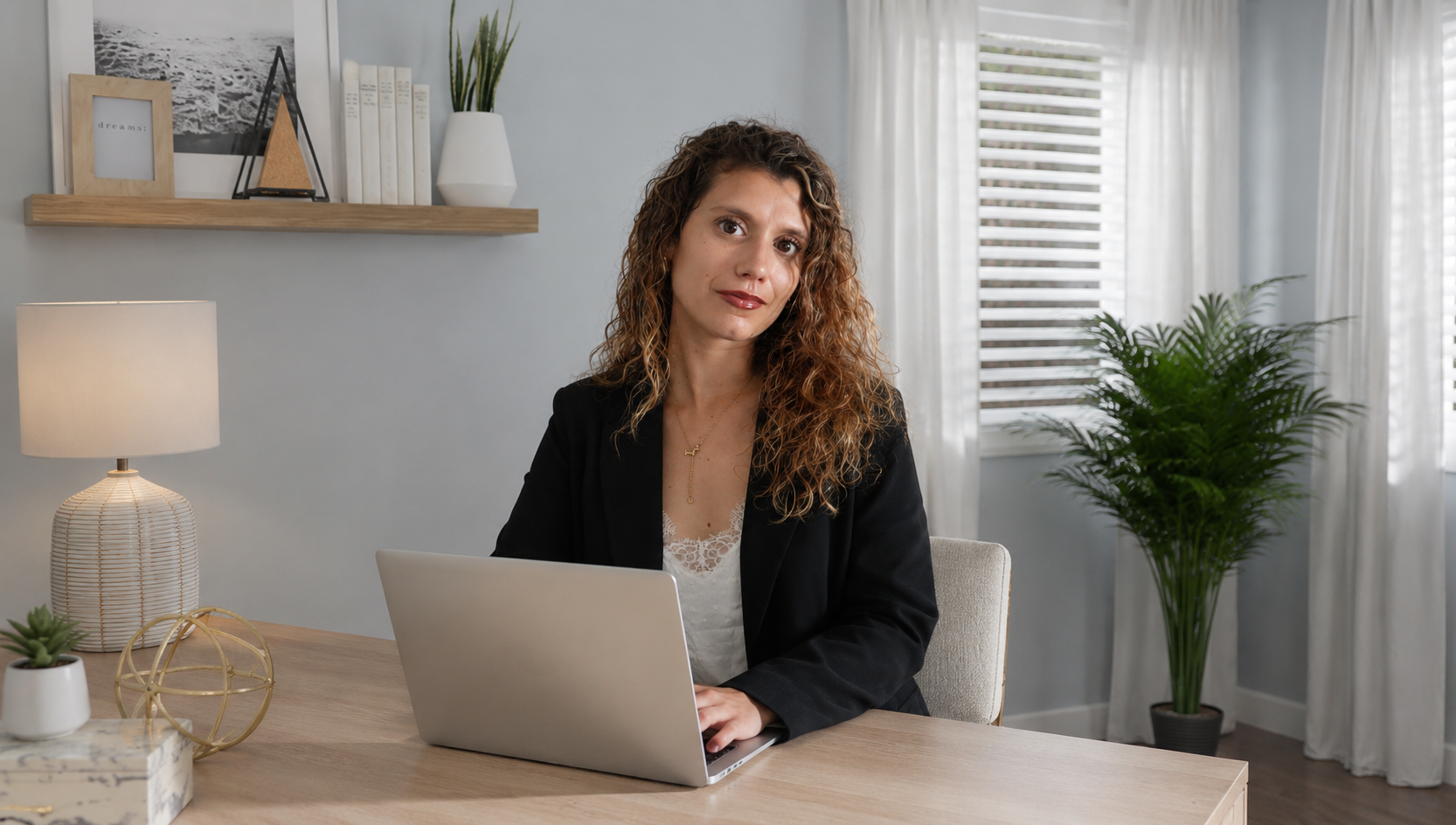 Mujer con cabello rizado arriba de los hombros, sentada en una mesa de madera, con una laptop plateada y ropa formal negra, en un salón decorado con plantas y cuadros en las paredes, iluminado con luz natural y lámpara.