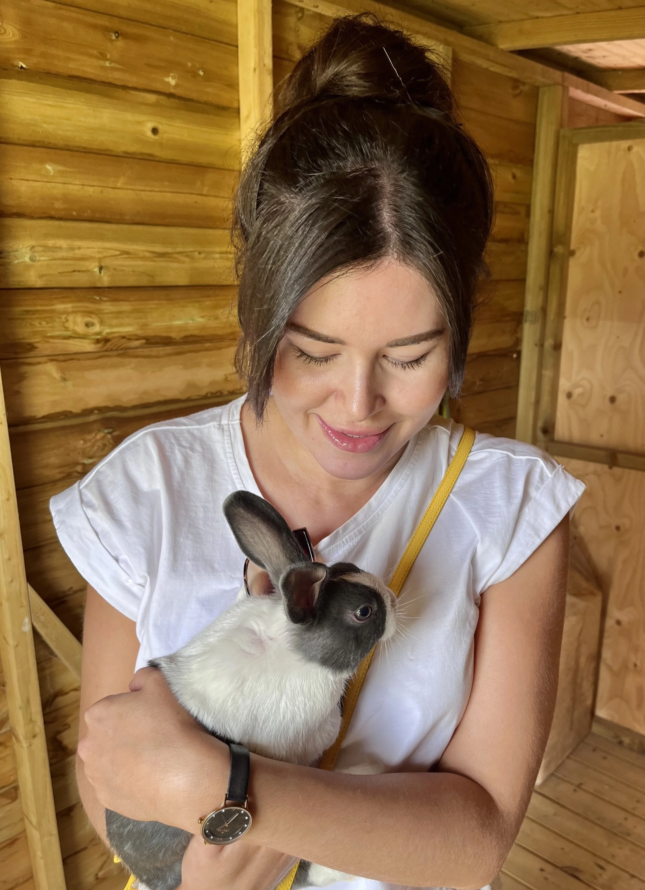 A woman with dark hair styled in an updo smiling and holding a small rabbit with black ears and black patches over the eyes, in a wooden room.