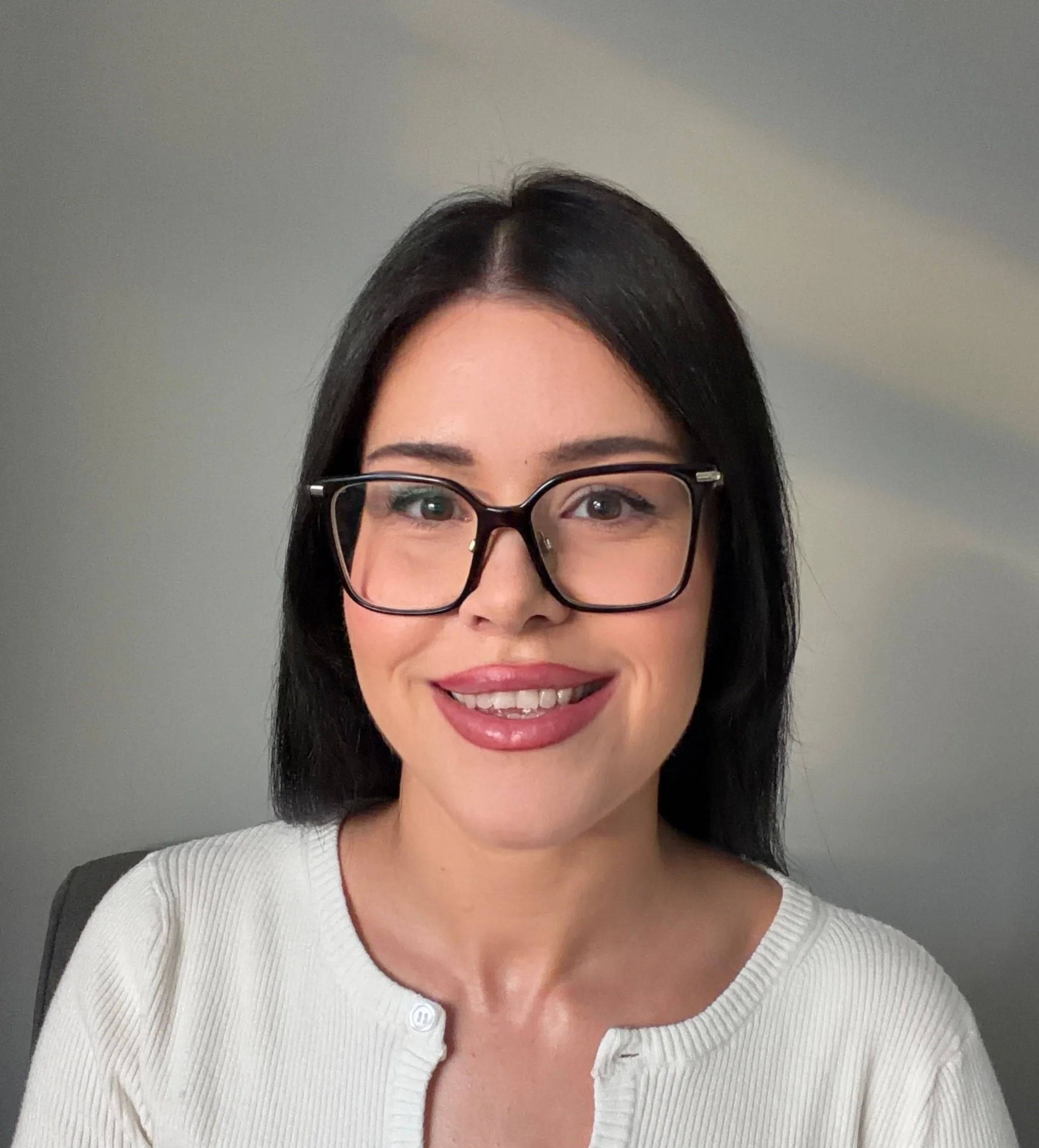 Close-up of a smiling woman wearing glasses and a white ribbed sweater, with dark hair and light skin, against a neutral background.