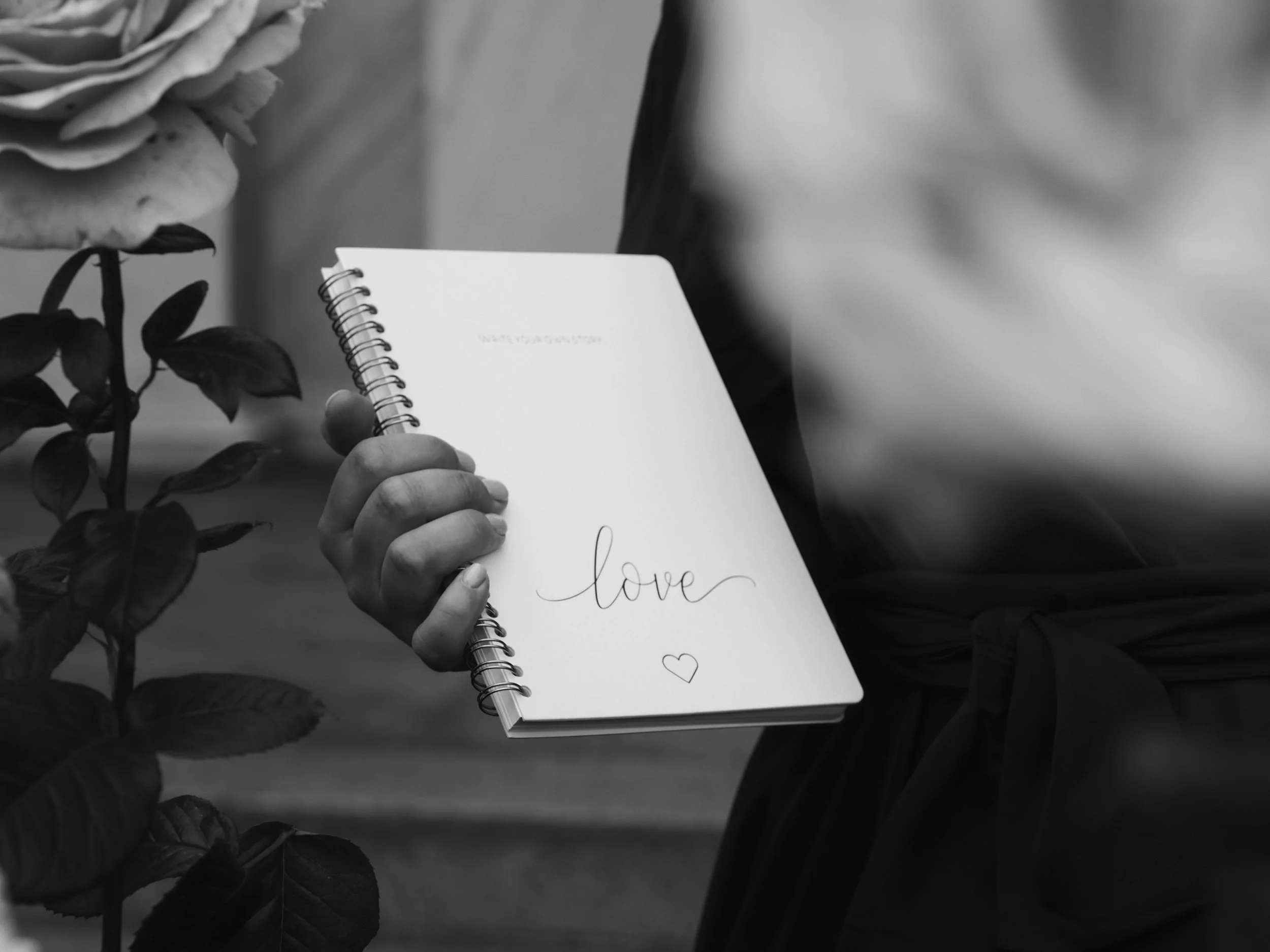 Eine Hand hält ein Notizbuch mit der Aufschrift ‚love‘ und einem kleinen Herz darunter. Auf der linken Seite sind Blätter einer Pflanze zu sehen.