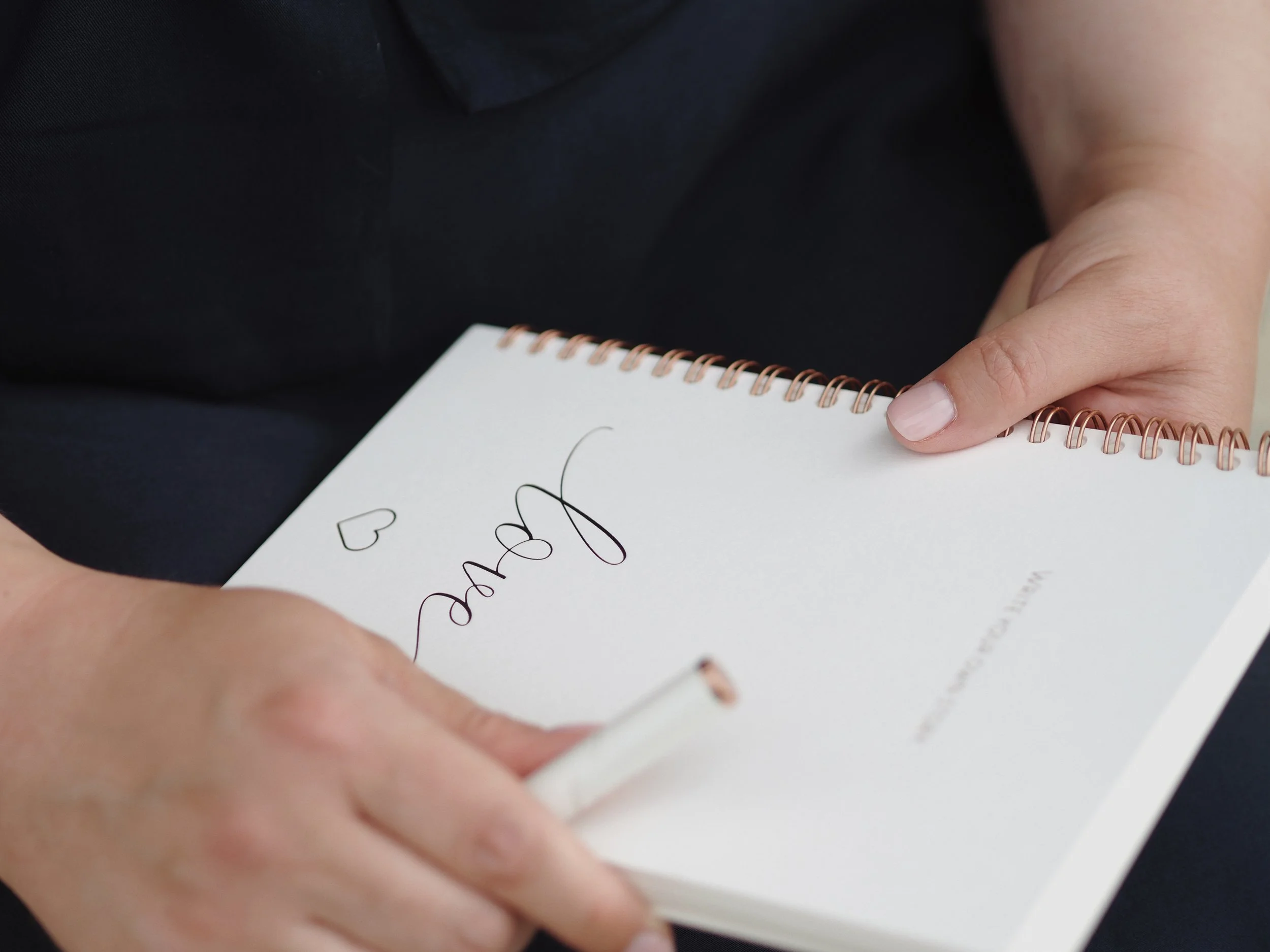 Eine Hand hält ein Notizbuch mit spiralförmigem Kupferrahmen, auf dem handschriftlich geschrieben steht, während im Hintergrund eine Person sitzt, die schwarze Kleidung trägt.