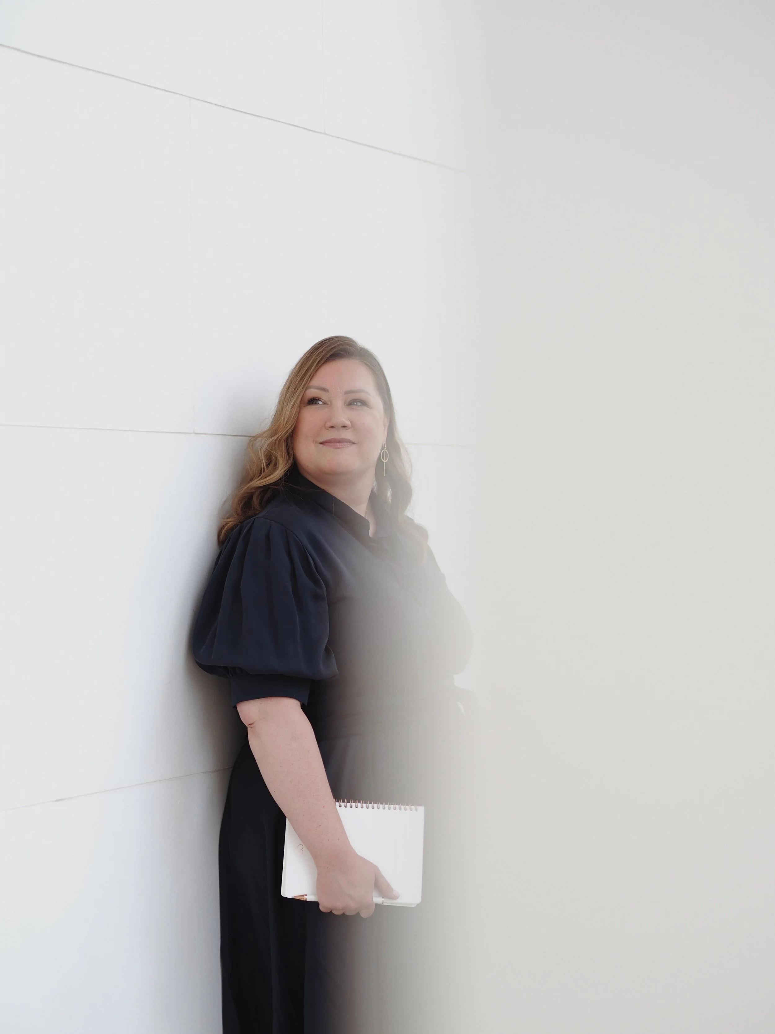 Eine Frau im schwarzen Kleid lehnt an einer weißen Wand, hält ein Notizbuch in der Hand und lächelt leicht.