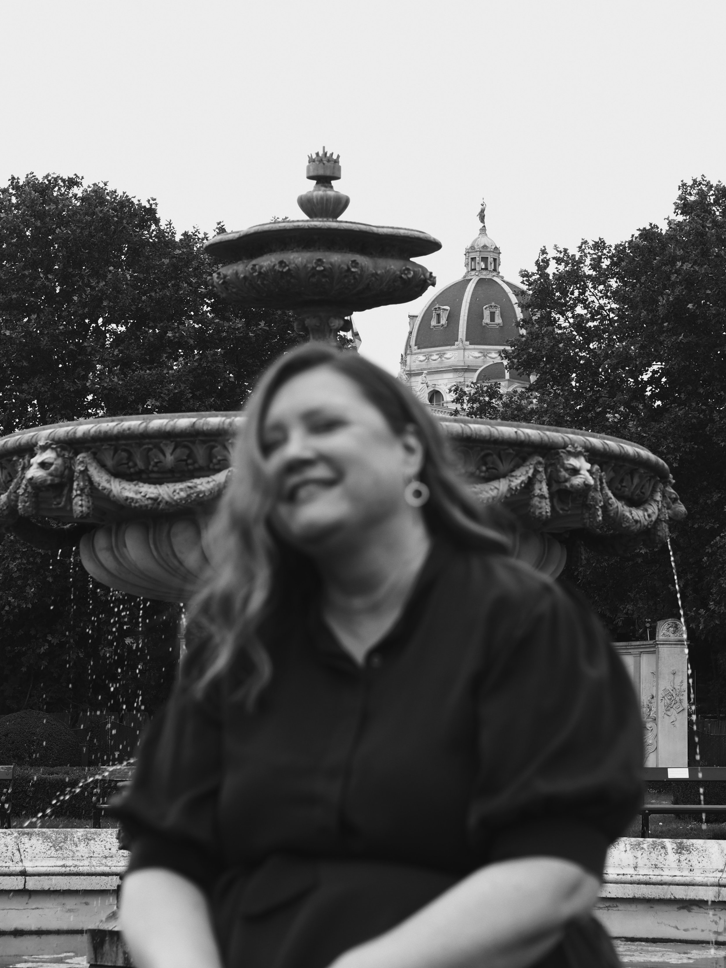 Schwarz-Weiß-Bild eines Brunnen mit einer Statue, im Hintergrund eine barocke Kuppel und umgeben von Bäumen.