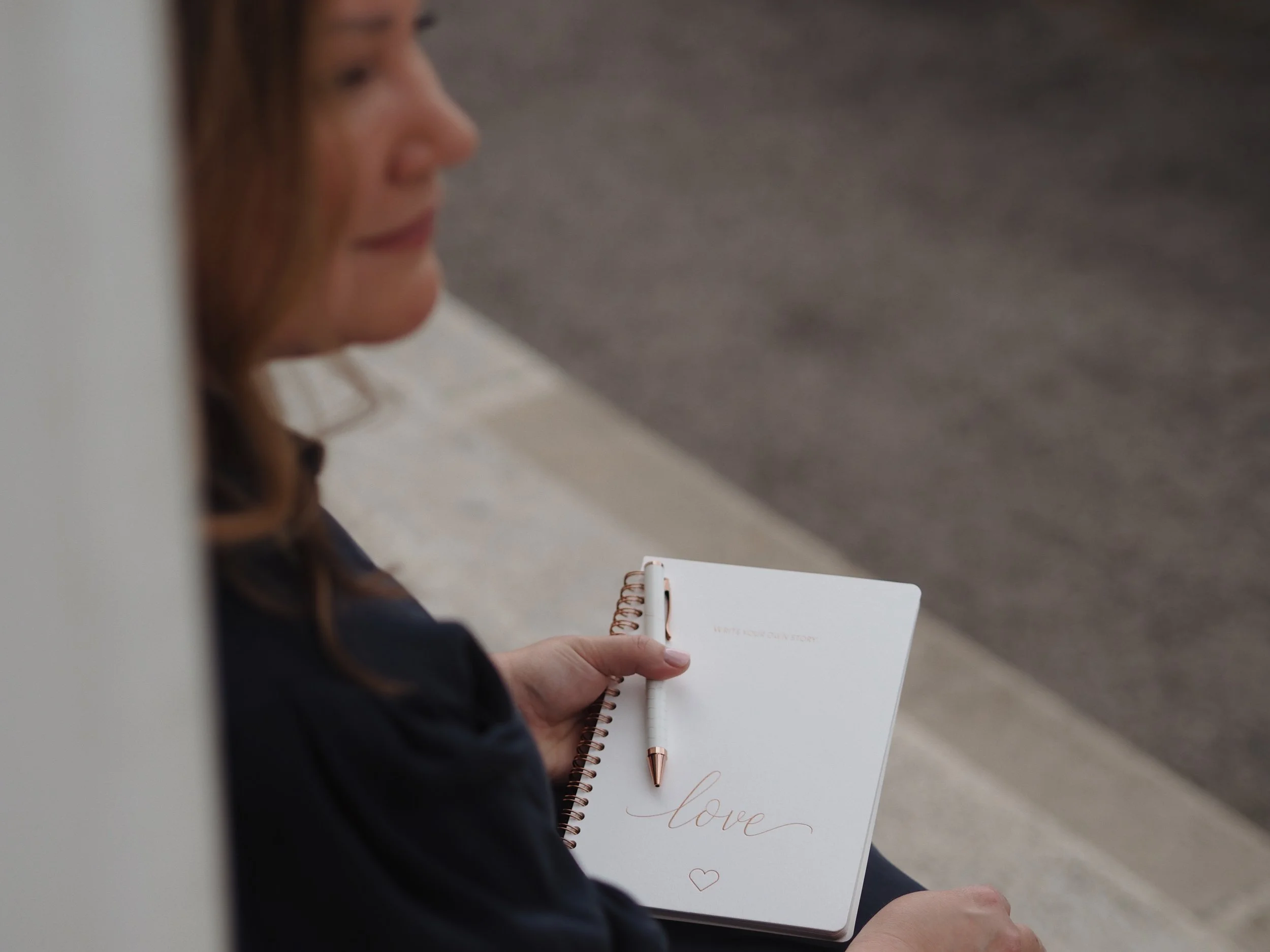 Eine Person sitzt auf dem Boden und hält ein offenes Notizbuch mit dem Wort "Liebe" und einem Herz darauf. Die Person trägt eine dunkle Jacke und hat braune, leicht gewellte Haare.